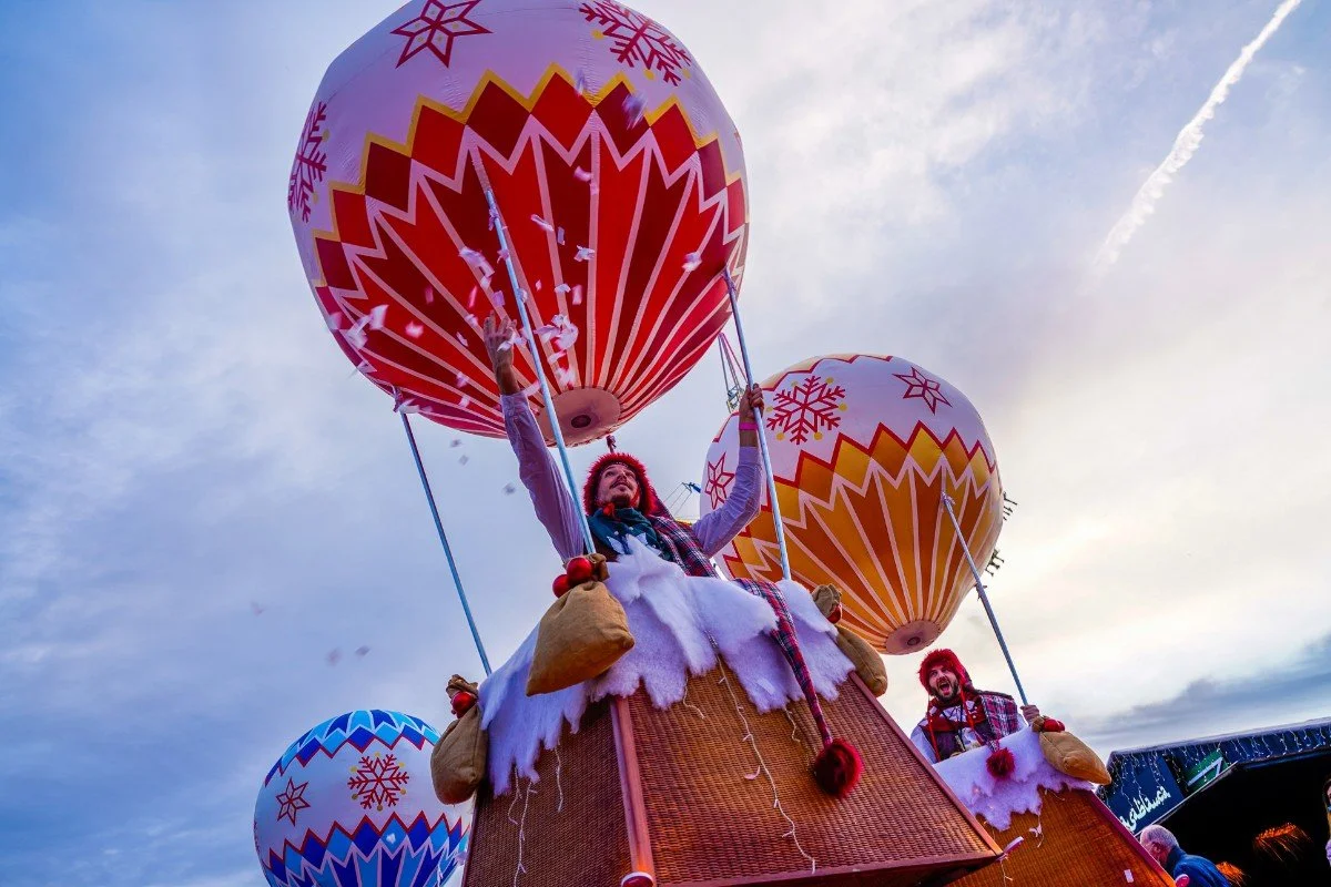 Personne dans une montgolfière décorée pour célébrer Noël, portant un costume festif, avec un ciel nuageux en arrière-plan.