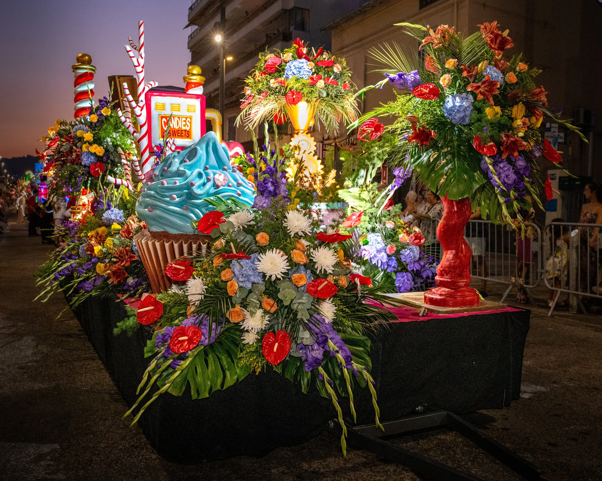 Un grand arrangement floral coloré, composé de fleurs rouges, violettes, jaunes et blanches, est disposé sur un socle noir. À l'arrière-plan, on aperçoit une enseigne « Bonbons », des bâtiments et des lampadaires.