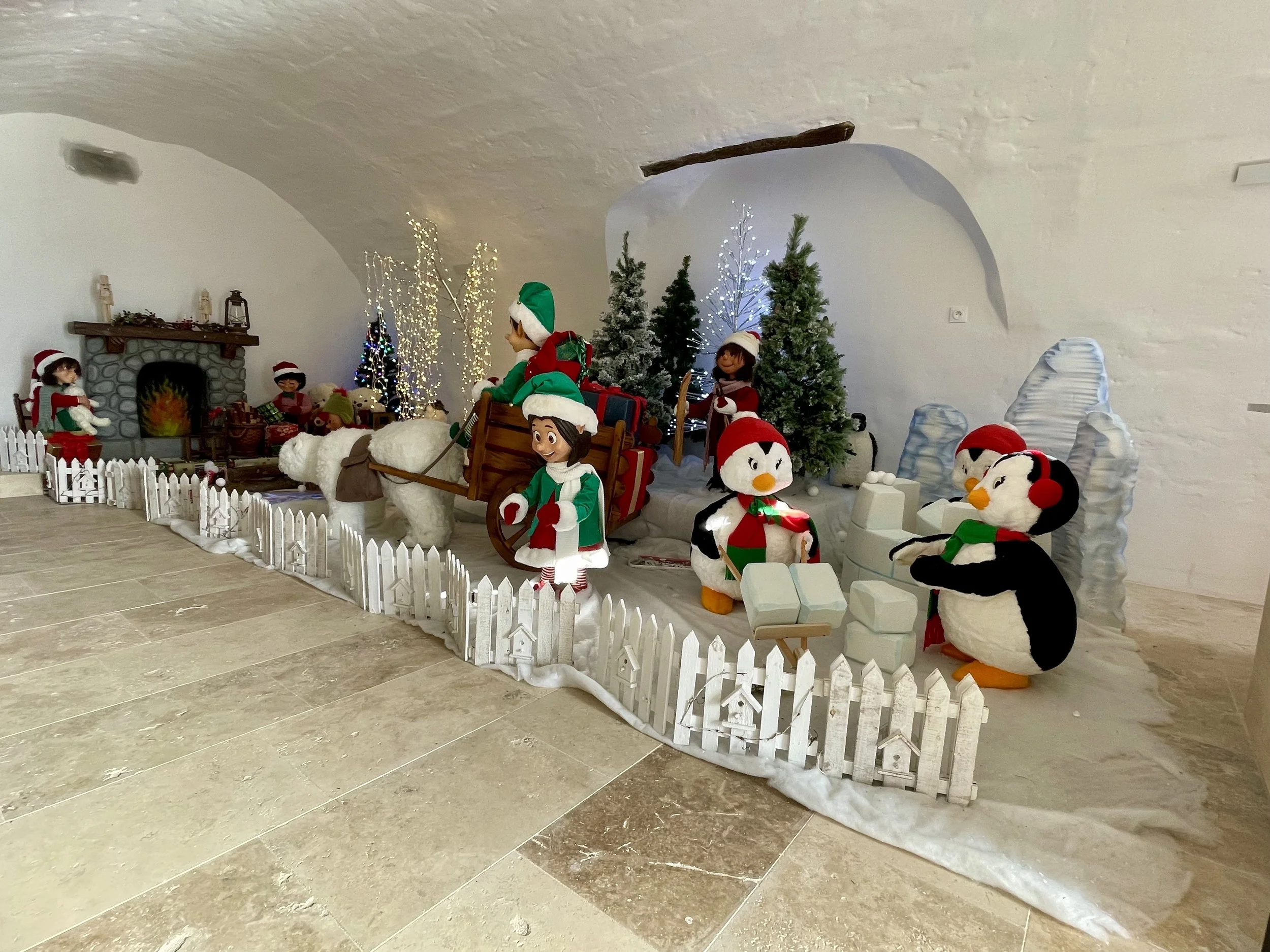 Décor de Noël avec des animaux en peluche et des lutins, des sapins de Noël décorés, une fausse cheminée et des découpages de paysages enneigés.