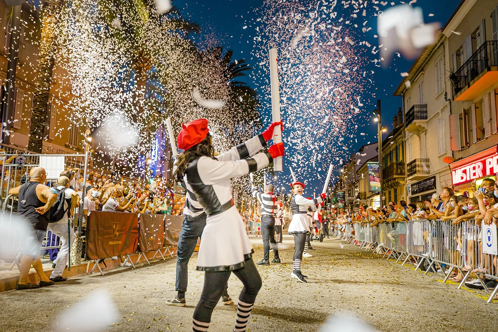 Une parade en soirée avec des participants en costumes de cirque, lançant des confettis dans la rue animée, entourée par un public qui regarde derrière des barrières.