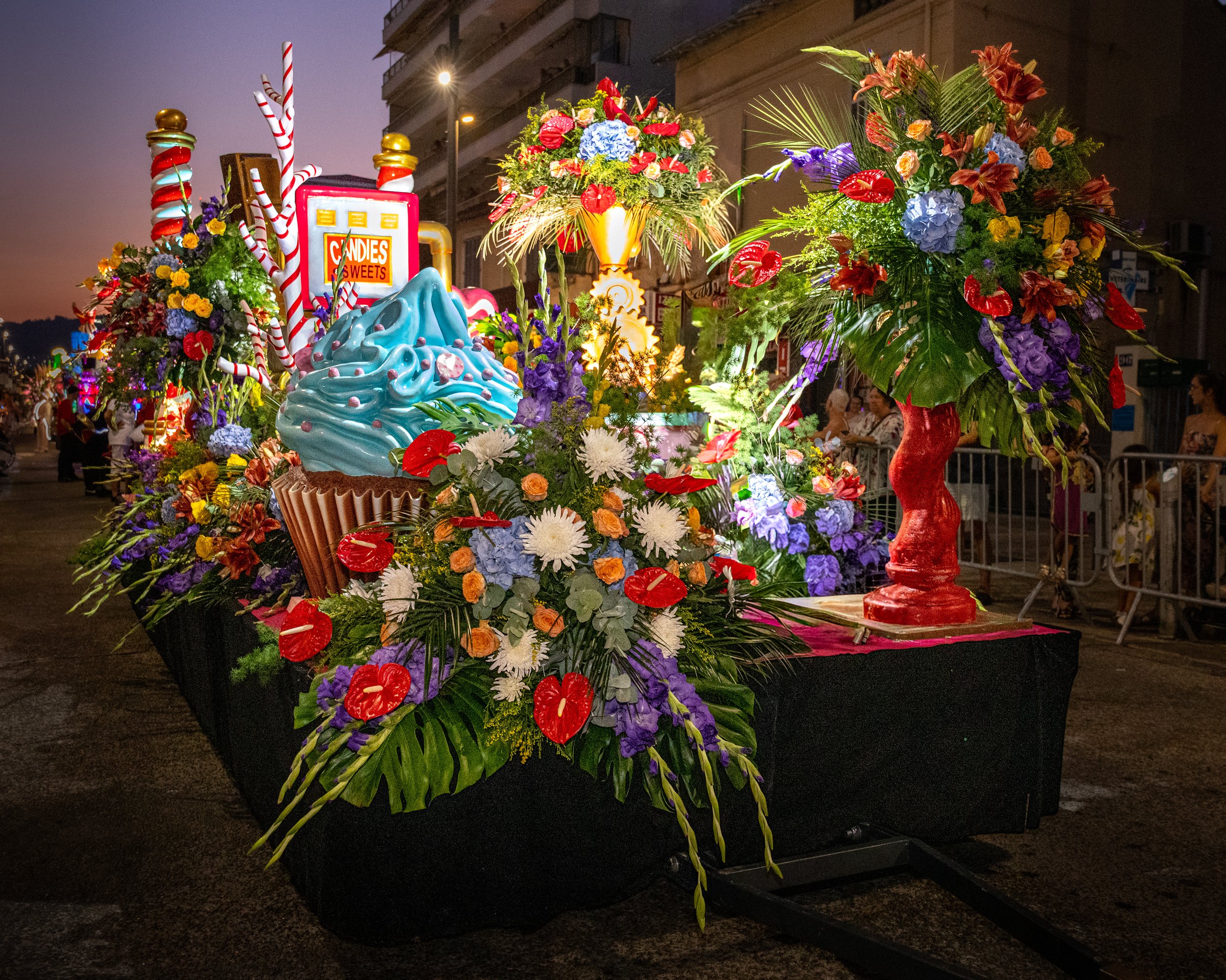 Composition florale éclatante sur un piédestal noir au crépuscule, présentant un assortiment de fleurs colorées, notamment rouges, bleues, violettes et jaunes, avec une pancarte décorative indiquant « CANDIES SWEETS » en arrière-plan, le tout sur fon