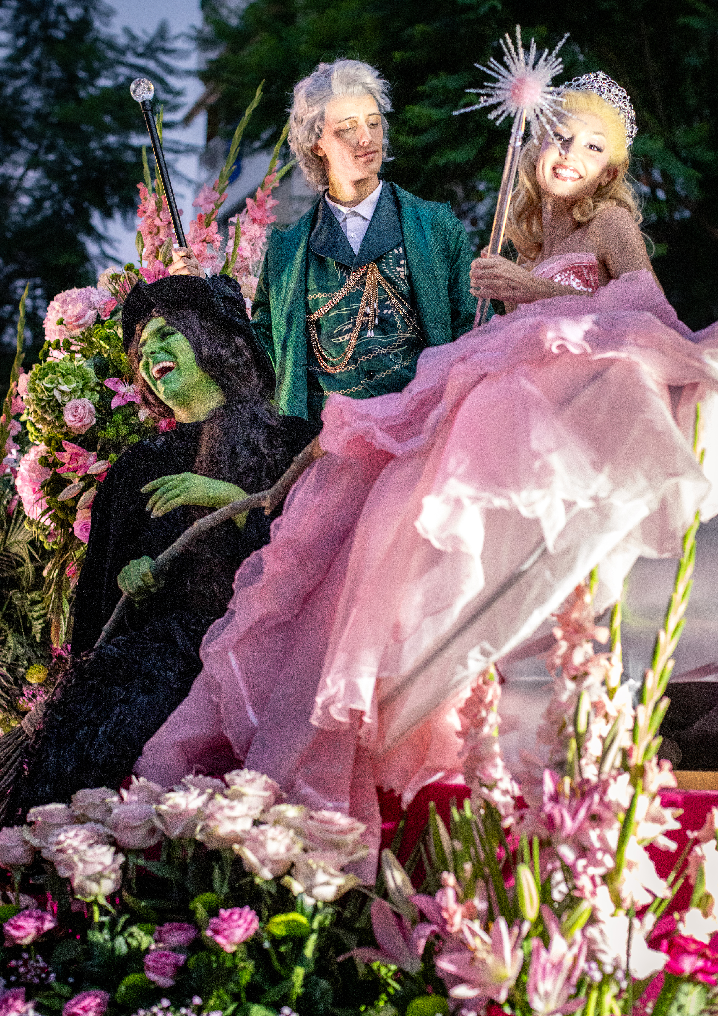 Personne déguisée en sorcière verte dans un décor floral, flanquée d’une femme en robe rose, entourée de fleurs dans un parc