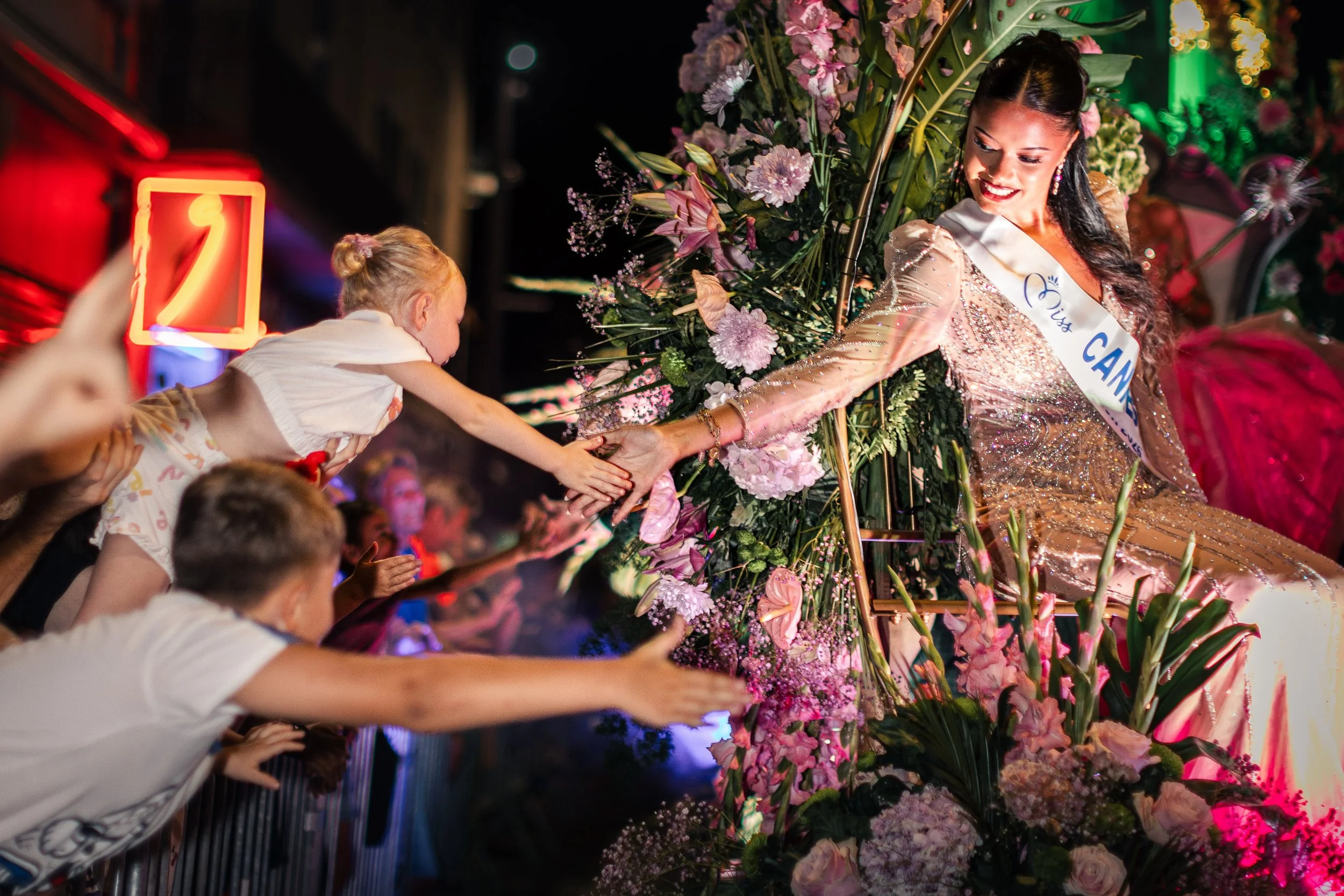 Un char allégorique décoré de fleurs roses et violettes, représentant une femme portant une écharpe où l'on peut lire « Mademoiselle » et une robe scintillante, tendant la main vers une jeune fille dans la foule.