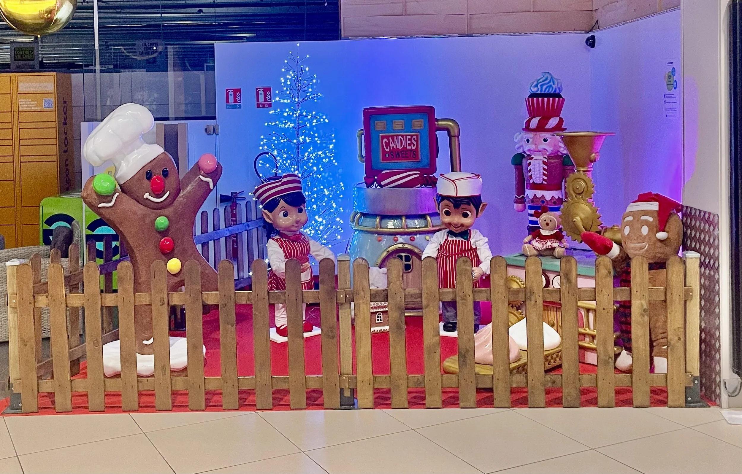 Décor de Noël avec des biscuits animés, des lutins, un sapin de Noël illuminé en bleu et une pancarte indiquant « Bonbons et Friandises », le tout derrière une clôture en bois.