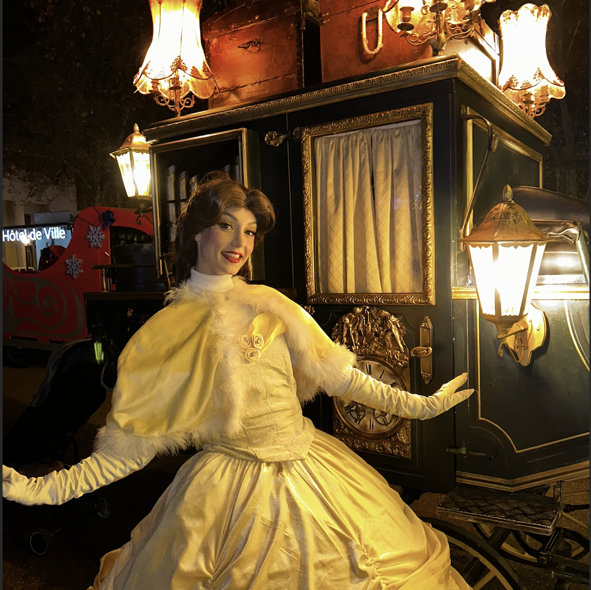Une femme vêtue d'une robe jaune élégante et d'une cape blanche avec une bordure en fourrure se tient devant un carrosse ancien illuminé par des lanternes.