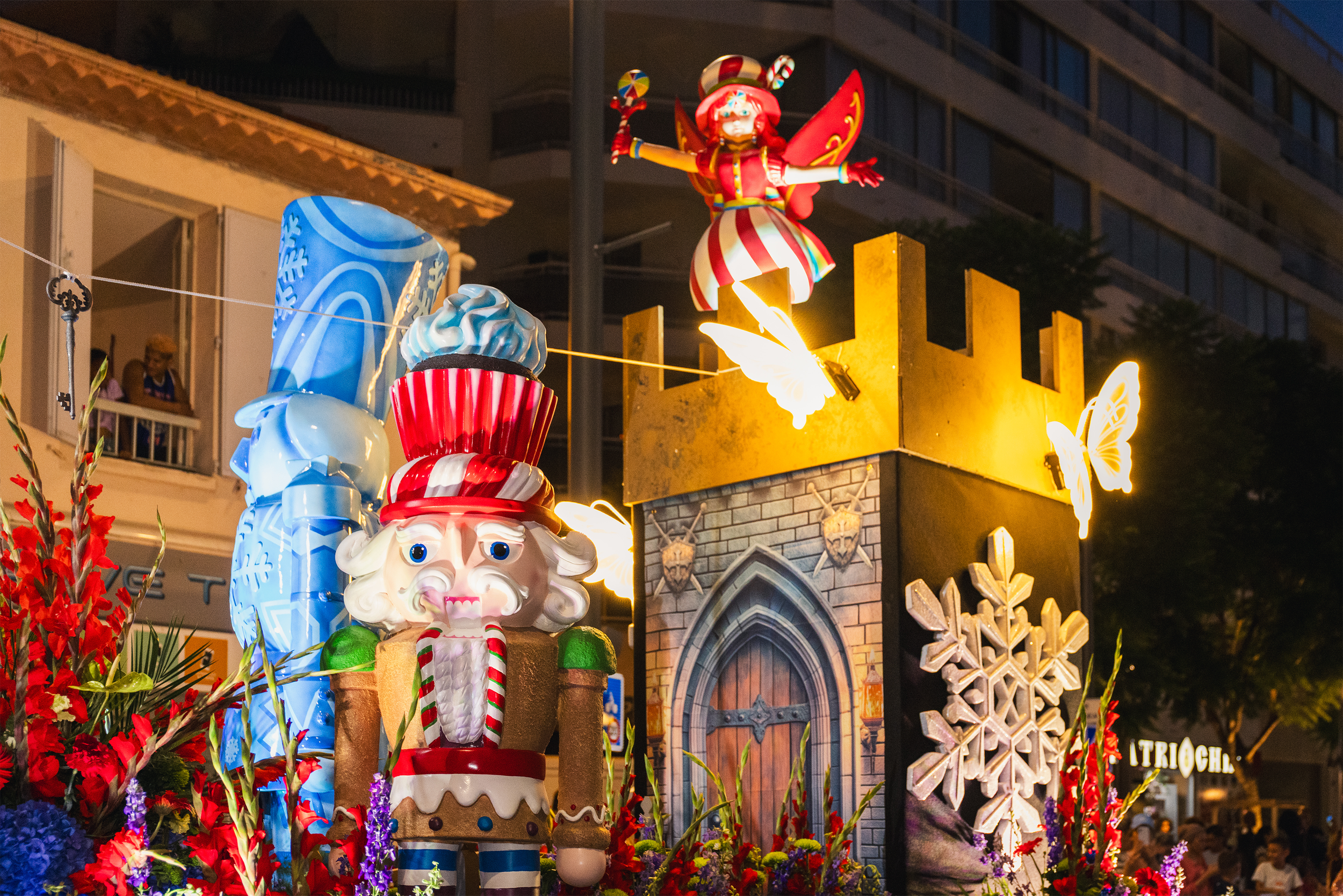 Décoration de fête avec un Père Noël, cupcakes, fleurs colorées, un bâtiment en arrière-plan et des lumières encadrant la scène.