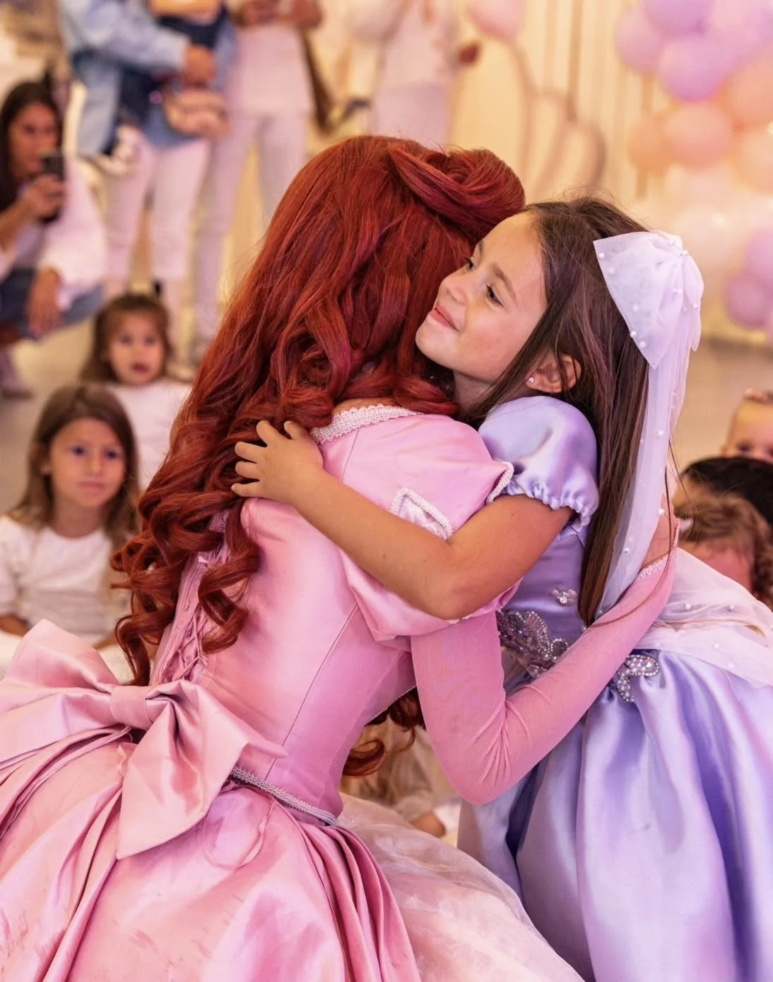 Une jeune fille en robe violette embrasse une femme en robe rose lors d'une célébration, entourée d'autres enfants et adultes en arrière-plan.