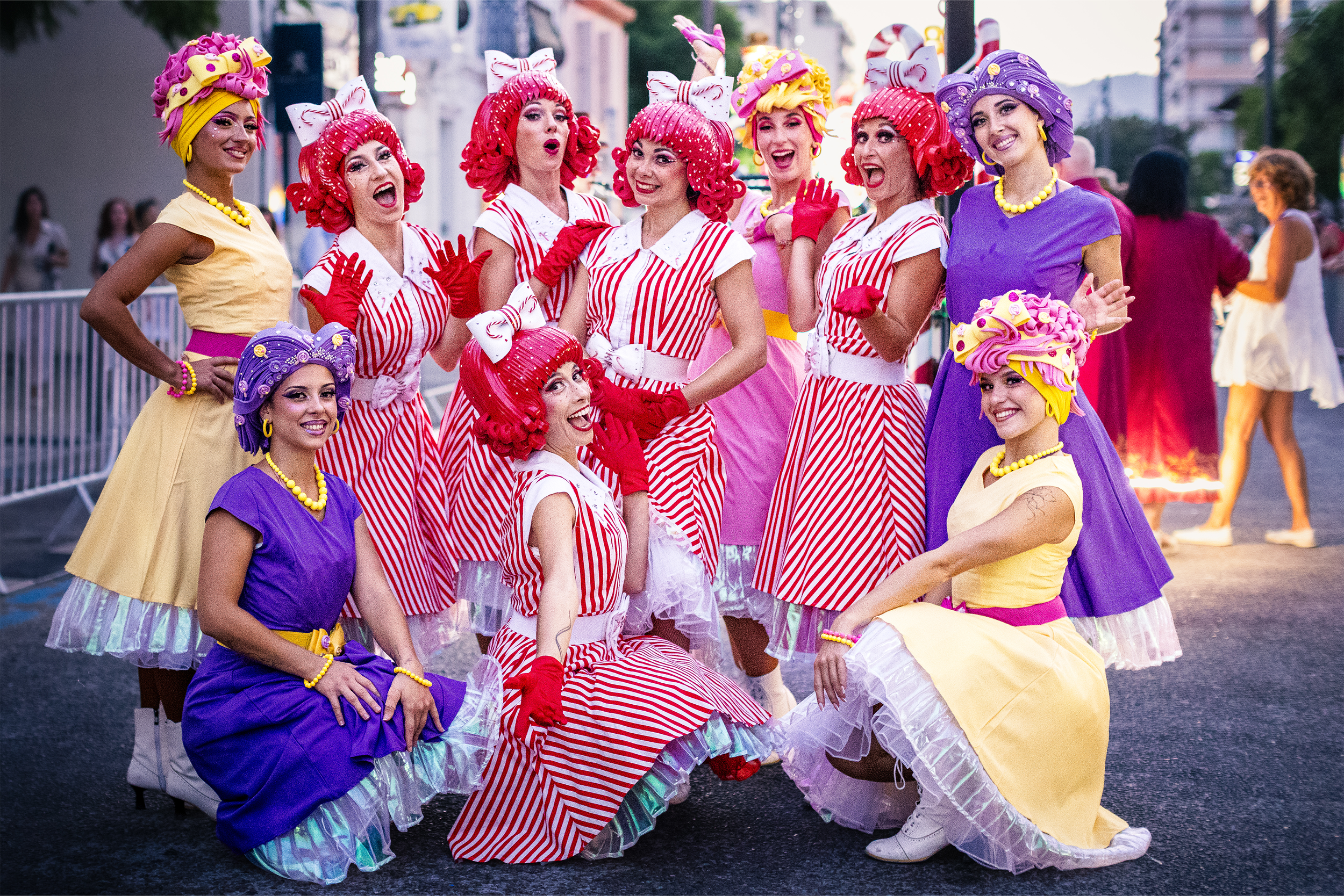 Groupe de femmes déguisées en personnages colorés avec des robes à rayures rouges et blanches, des accessoires de maquillage éclatants et des coiffures extravagantes, posant et souriant lors d'un festival ou d'une parade en plein air.