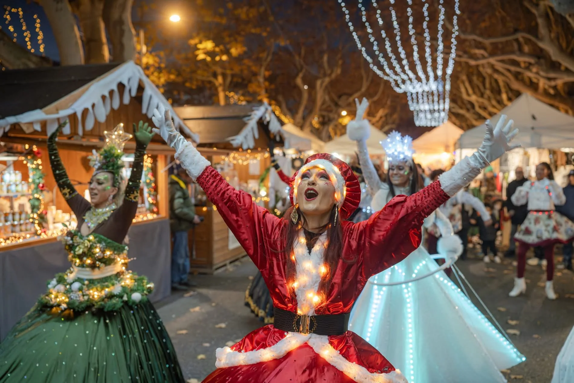 Des personnes déguisées en personnages de Noël dans un marché de Noël en soirée, avec des stands en bois décorés de lumières, et des arbres illuminés en arrière-plan. Dans les alpes maritimes 06 à cannes