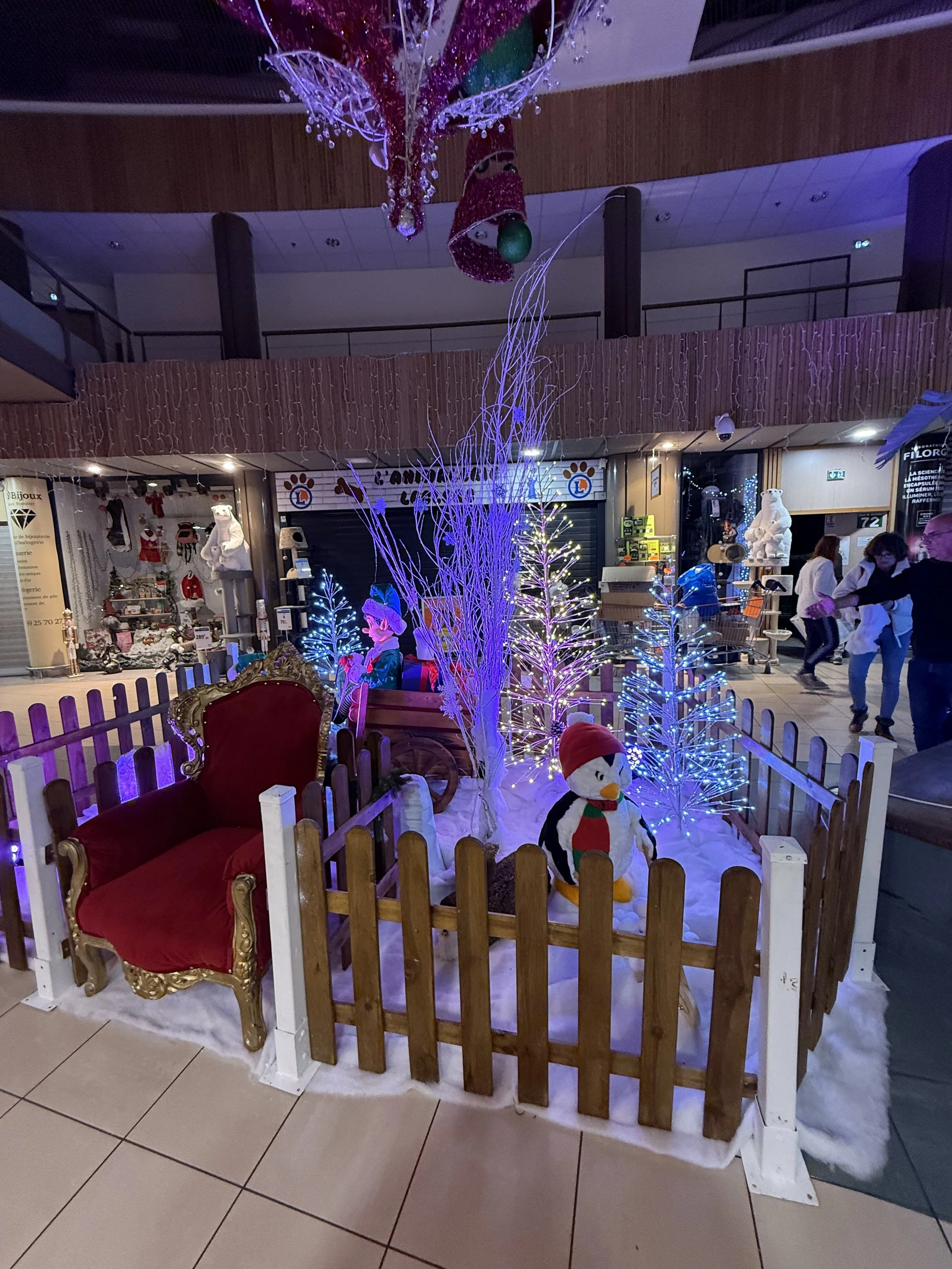 Décoration de Noël comprenant un trône du père noël en velours rouge à bordures dorées, une clôture blanche, un socle imitant la neige, un bonhomme de neige, des mini sapins de Noël bleus illuminés,