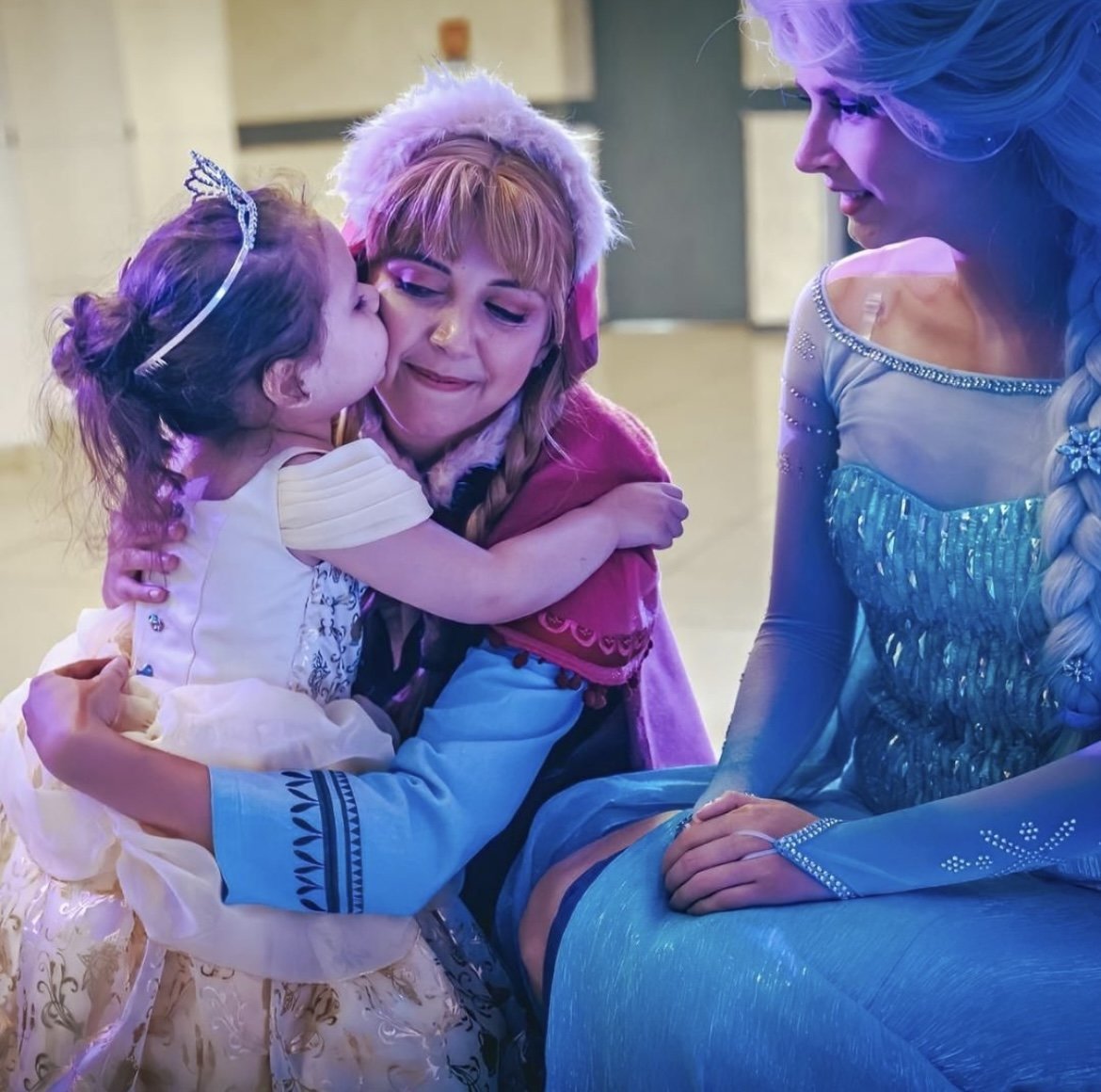 Une jeune fille embrasse une femme déguisée en Elsa de Disney, tandis qu'une autre femme déguisée en Elsa regarde avec un sourire, dans une scène colorée et joyeuse.
