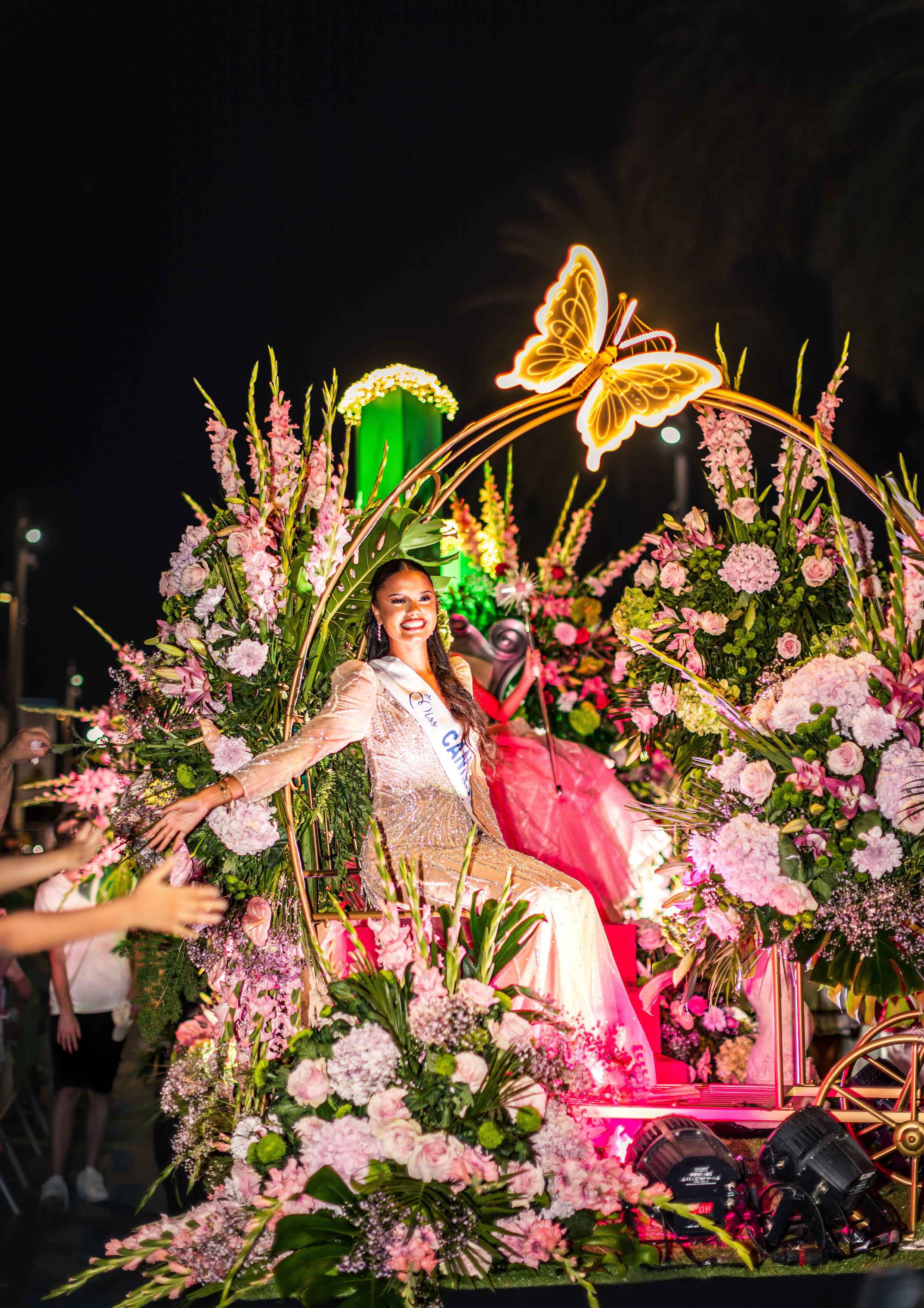Une femme portant une robe élégante assise sur un chariot décoré de nombreuses fleurs roses et vertes, dans un décor nocturne avec un papillon lumineux en façade et des lumières colorées en arrière-plan.