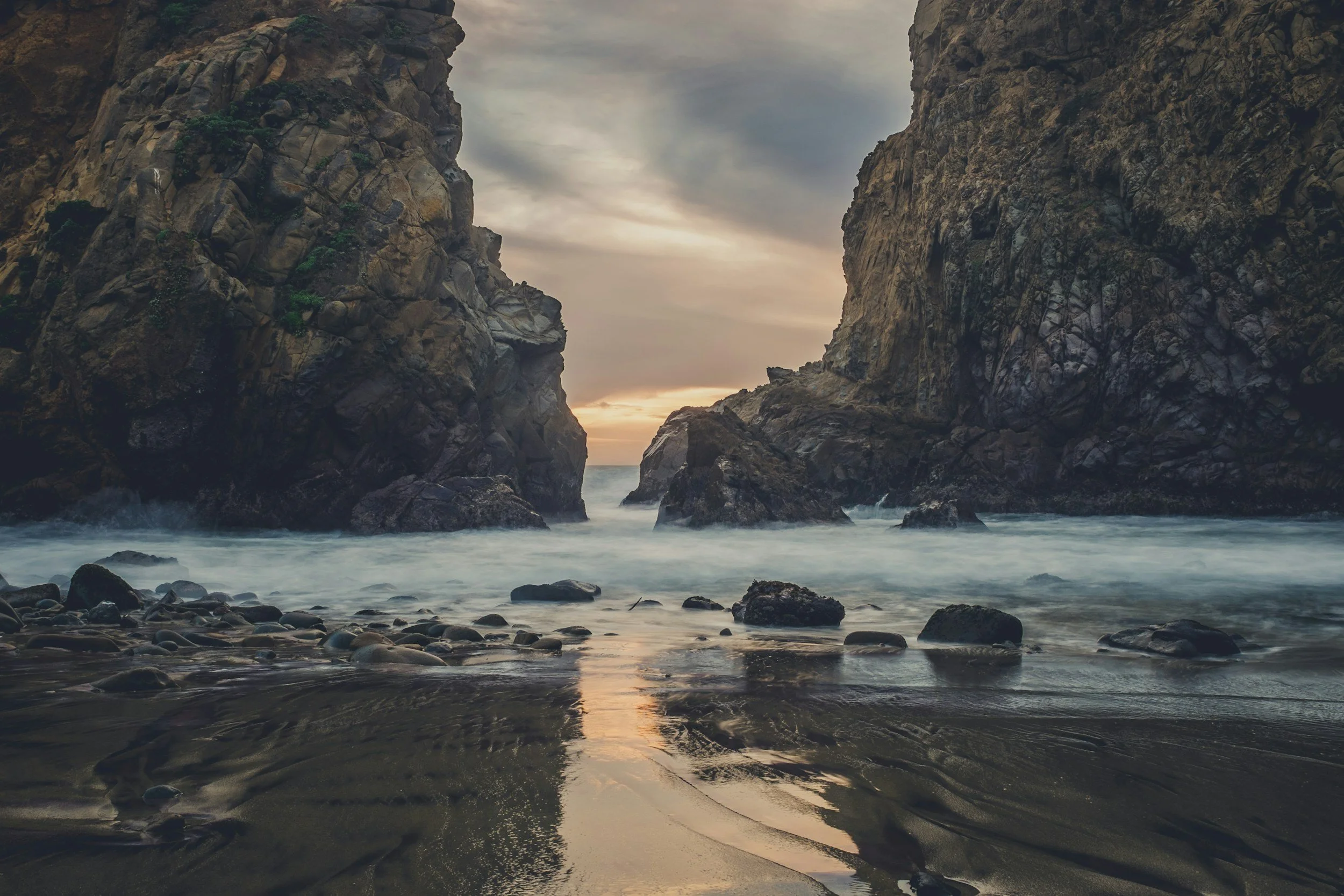 A scenic view of a narrow ocean inlet with high rocky cliffs on both sides during sunset, with rocks and pebbles on the shoreline and calm water reflecting the cloudy sky.