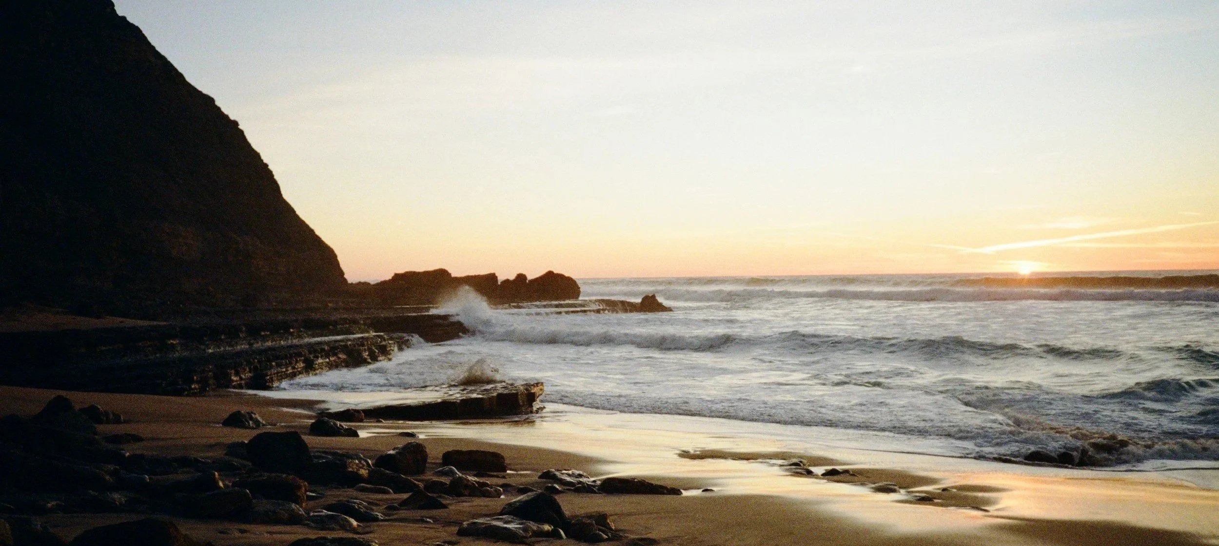 Sunset over a rocky ocean shoreline with waves crashing against rocks and a large cliff on the left side.
