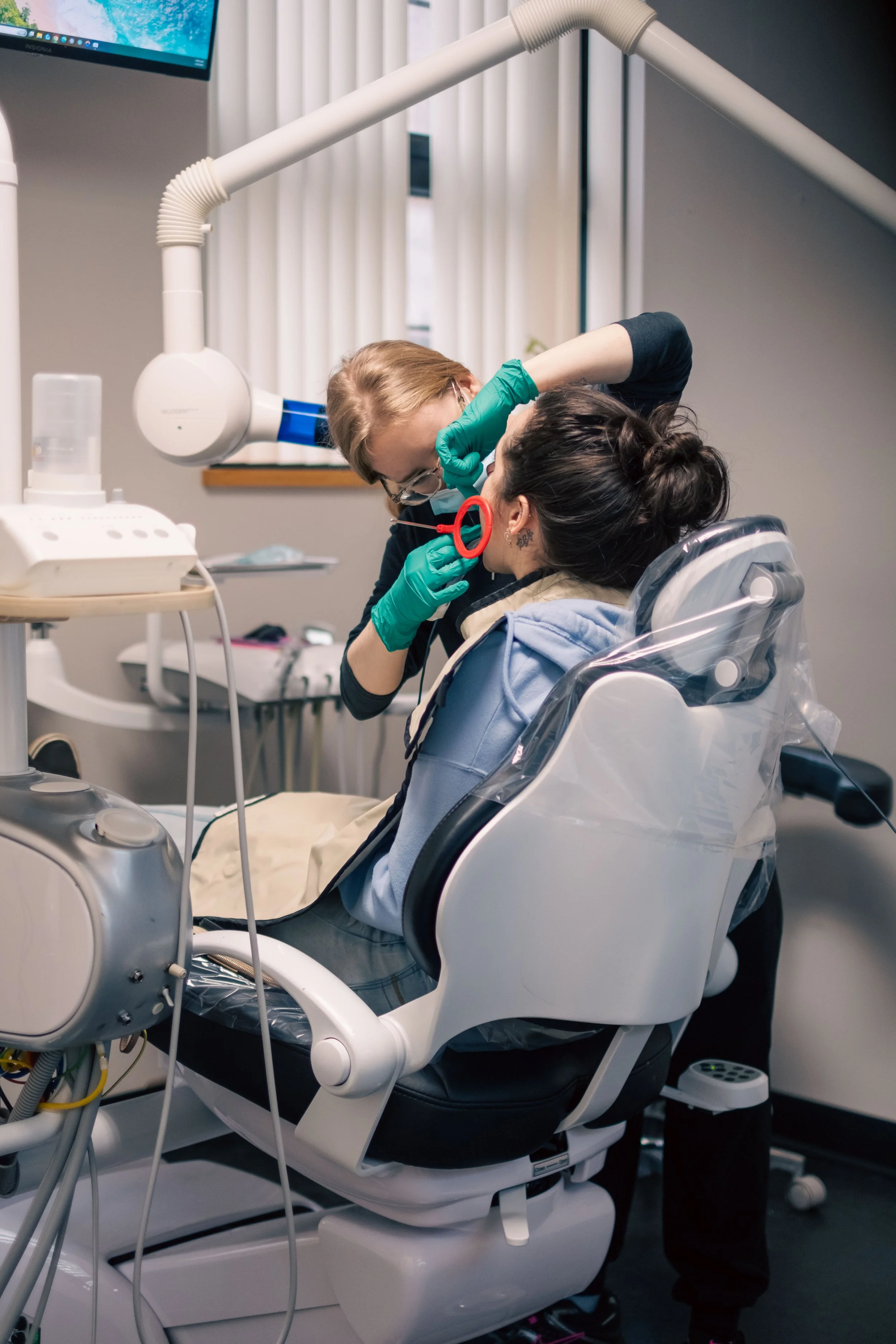 A dentist or dental hygienist examining a patient in a dental chair. The professional is wearing gloves and a mask, using dental tools to examine the patient's mouth. The setting is a modern dental clinic with equipment and a monitor visible in the background.