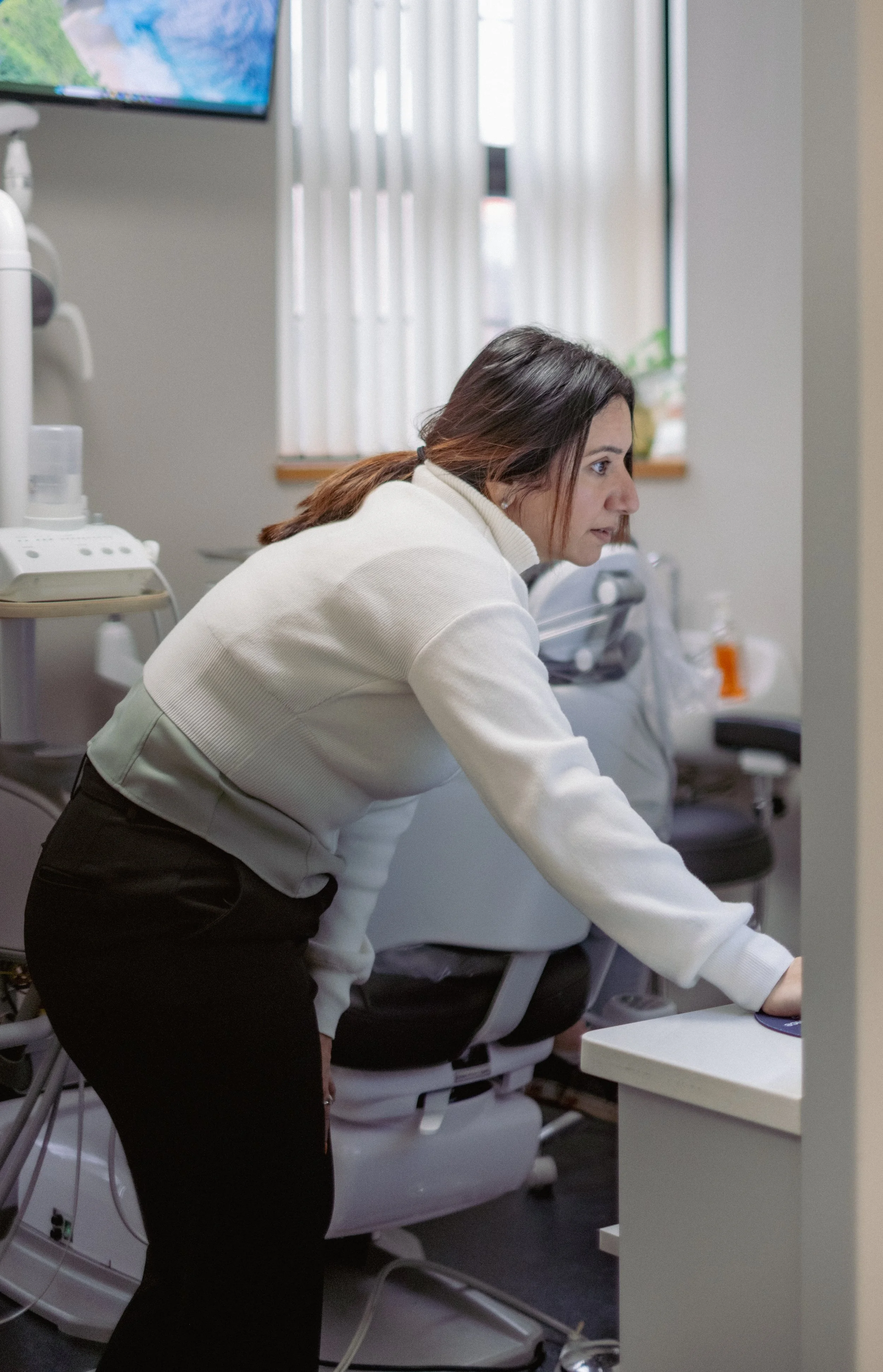 A woman with brown hair tied back, wearing a white sweater, leaning over a hospital bed with her right arm extended, possibly working on medical equipment or patient records in a hospital room with medical devices and a window with blinds.