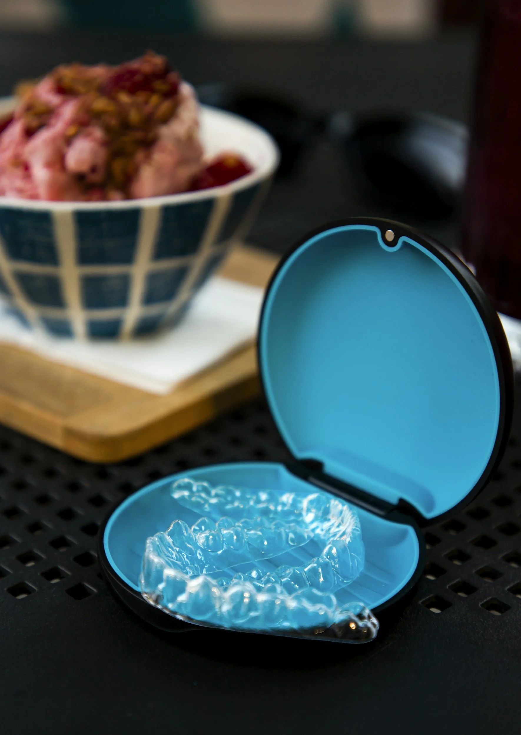 Open blue container with clear dental aligners inside, on a black surface. In the background, a bowl of mixed berries and a wooden cutting board are blurred.