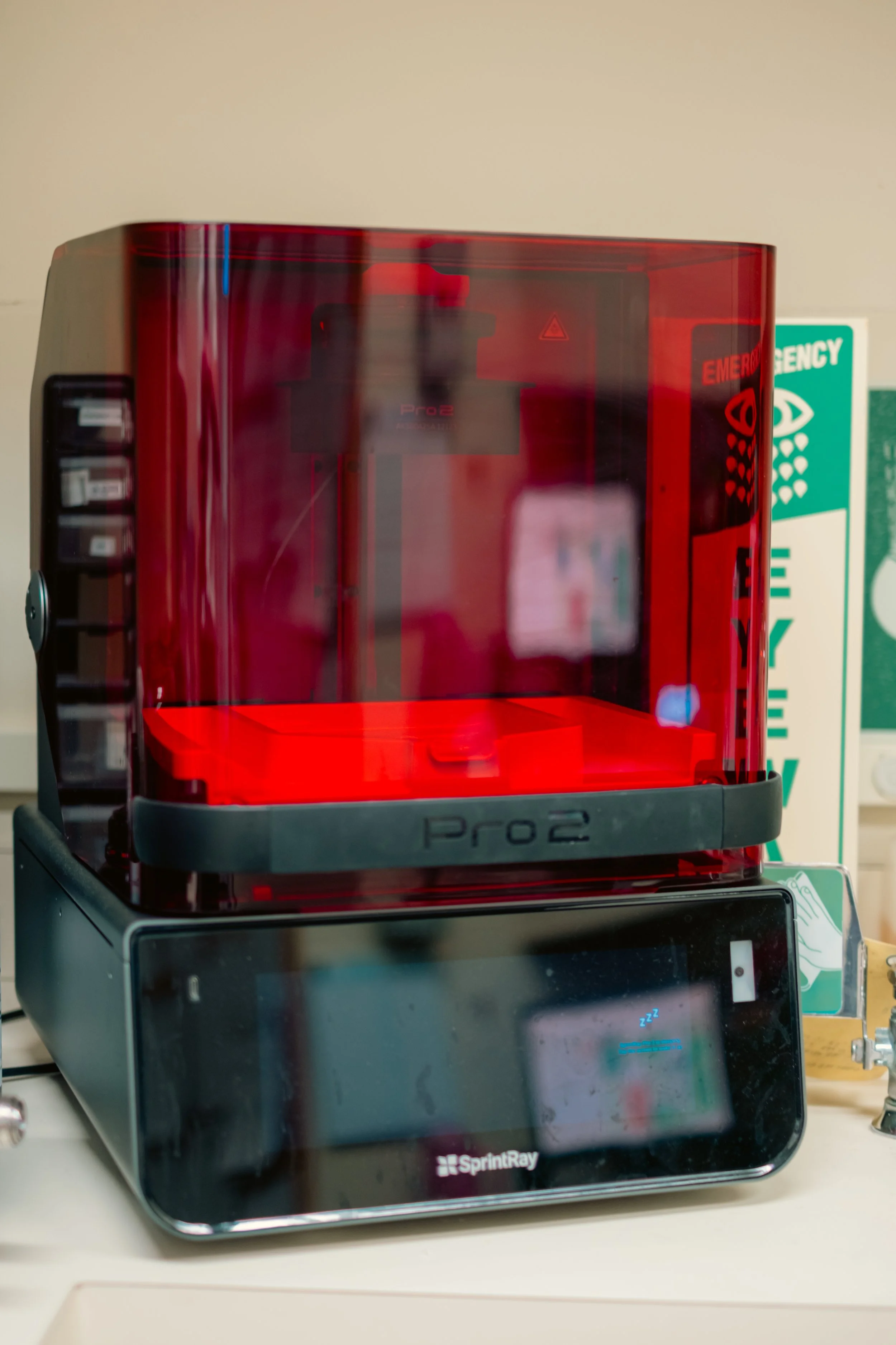 A red and black 3D printer on a desk, with a green emergency exit sign in the background.