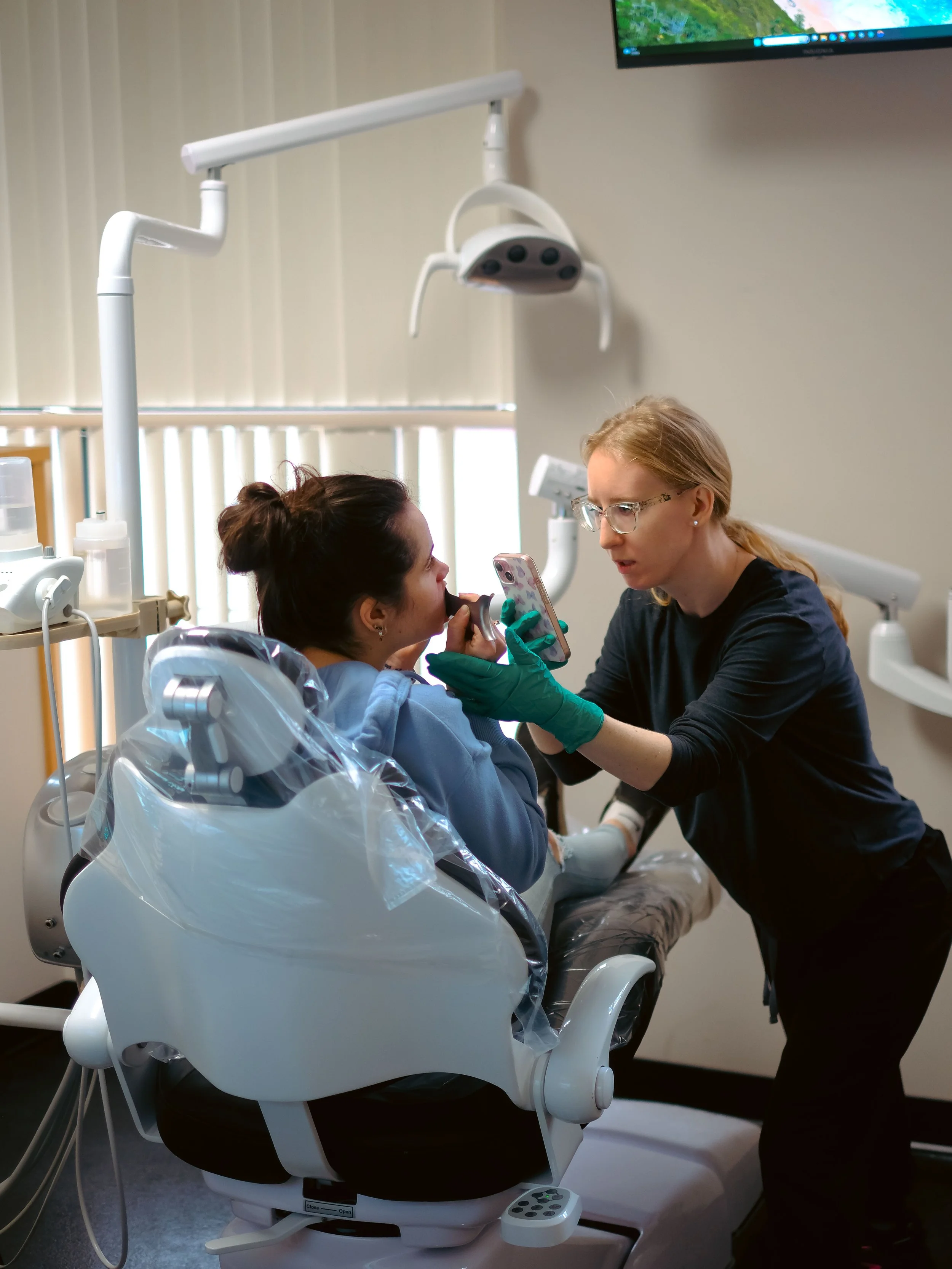 A healthcare professional is talking to a female patient in a hospital room. The patient is seated in a specialized medical chair, and the healthcare worker is holding a smartphone, possibly showing her information. Medical equipment and monitors are visible in the room.