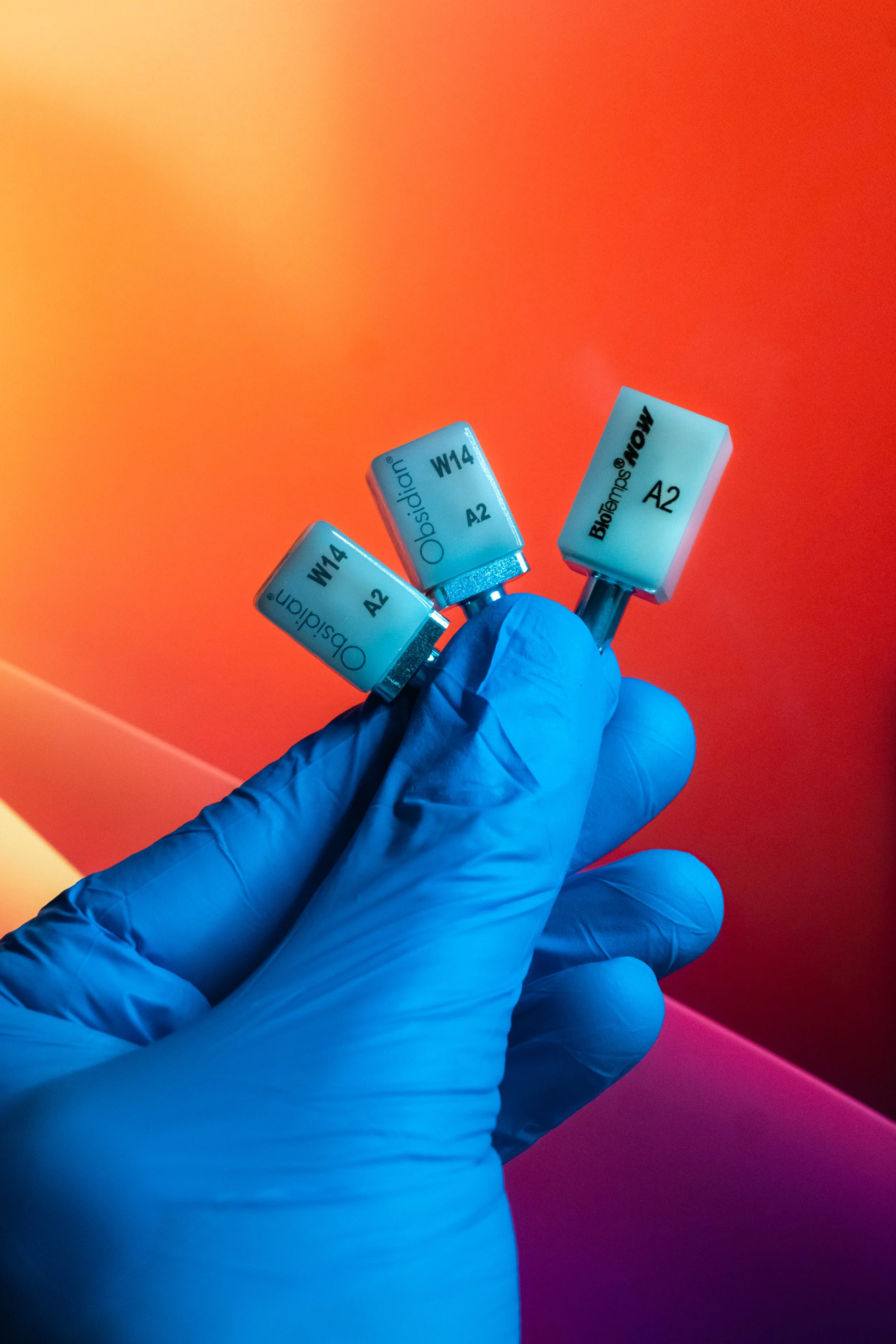 A hand wearing a blue glove holding three small bottles labeled 'Omnidip', 'Obsidian', and 'Bhotemps' with yellow and orange background.
