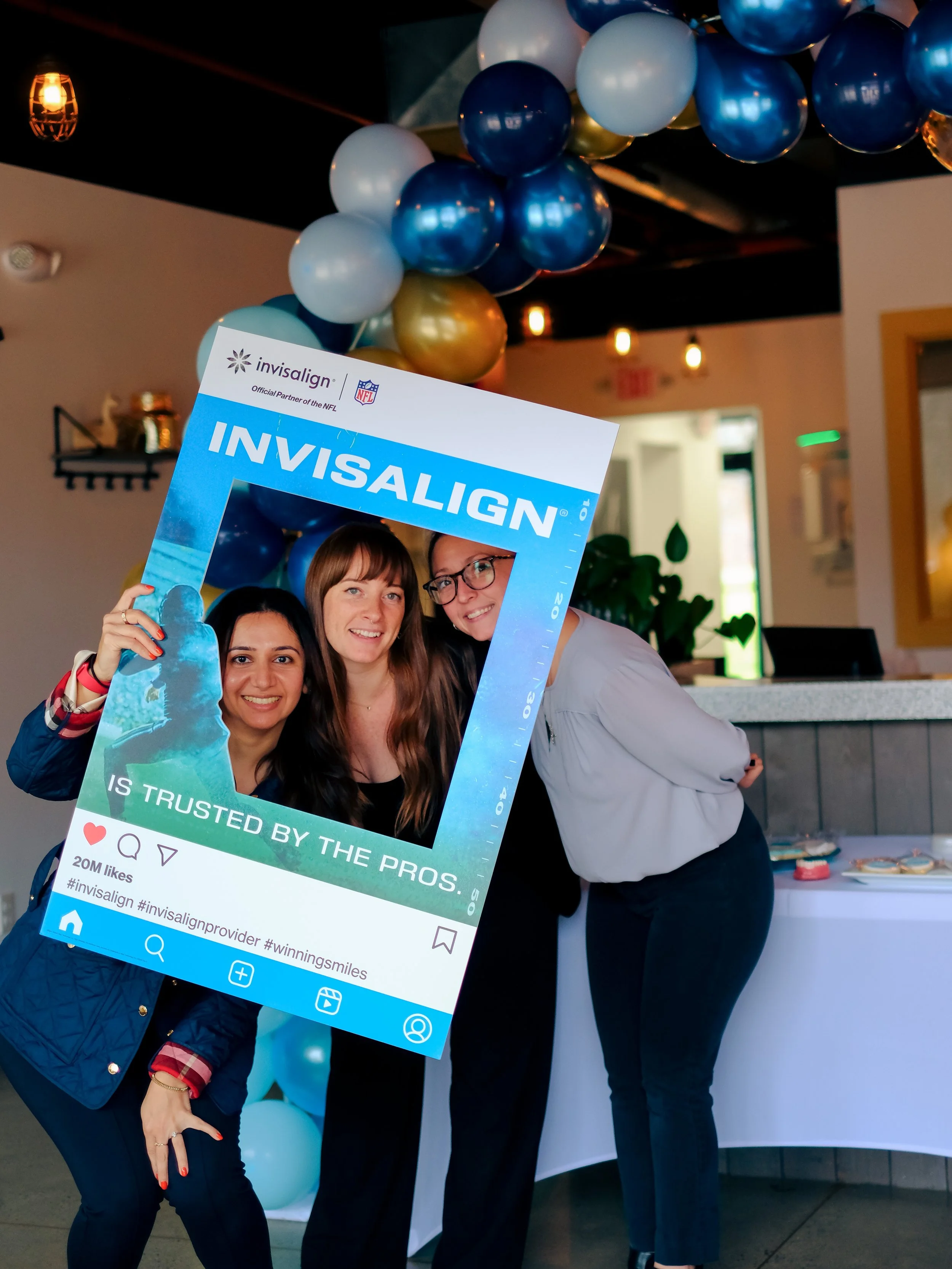 Three women holding a large inflatable social media frame at an indoor event, with blue, white, and gold balloons and warm lighting in the background.