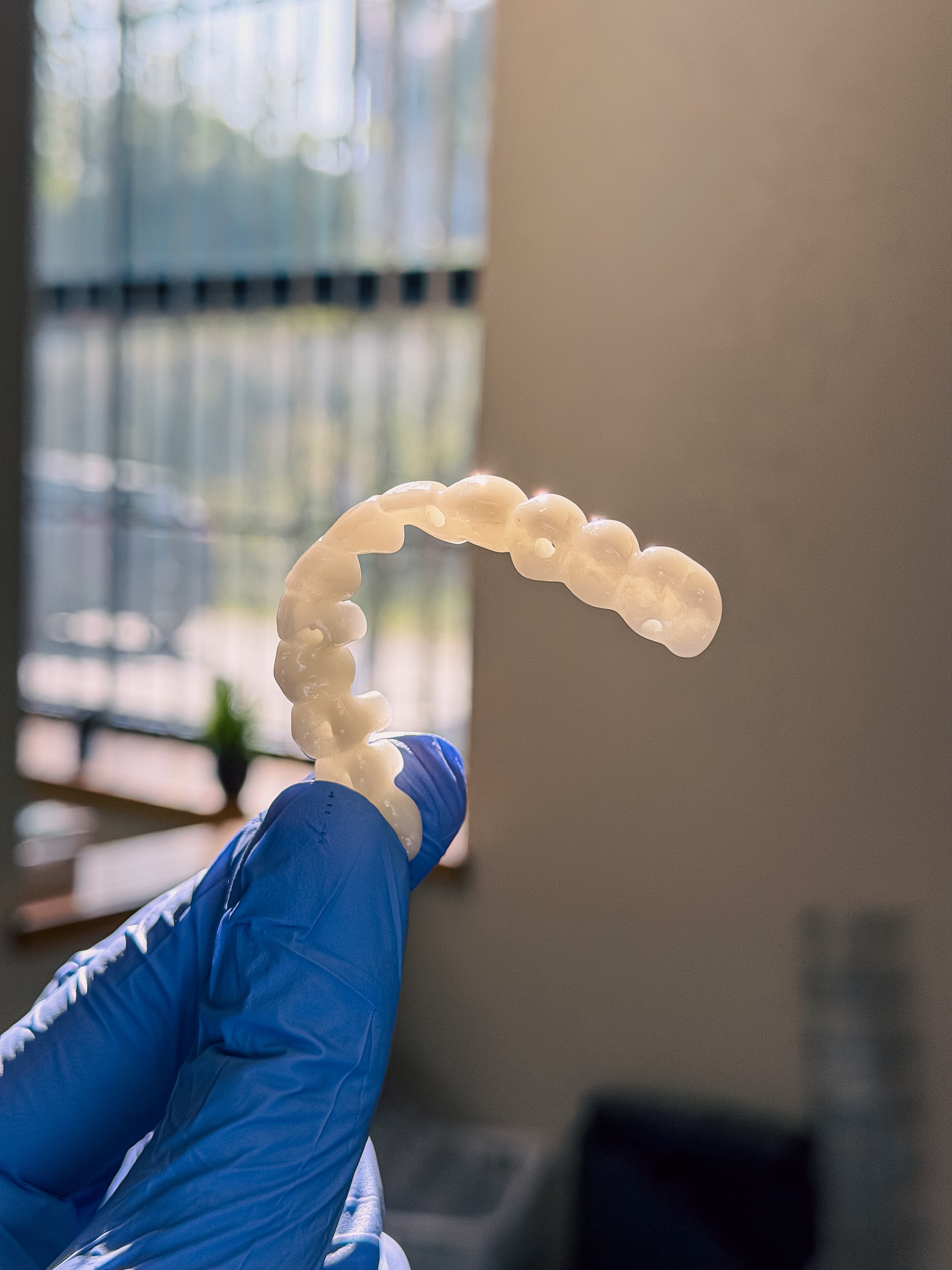 A person wearing a blue glove holding a dental aligner or retainer in front of a window.