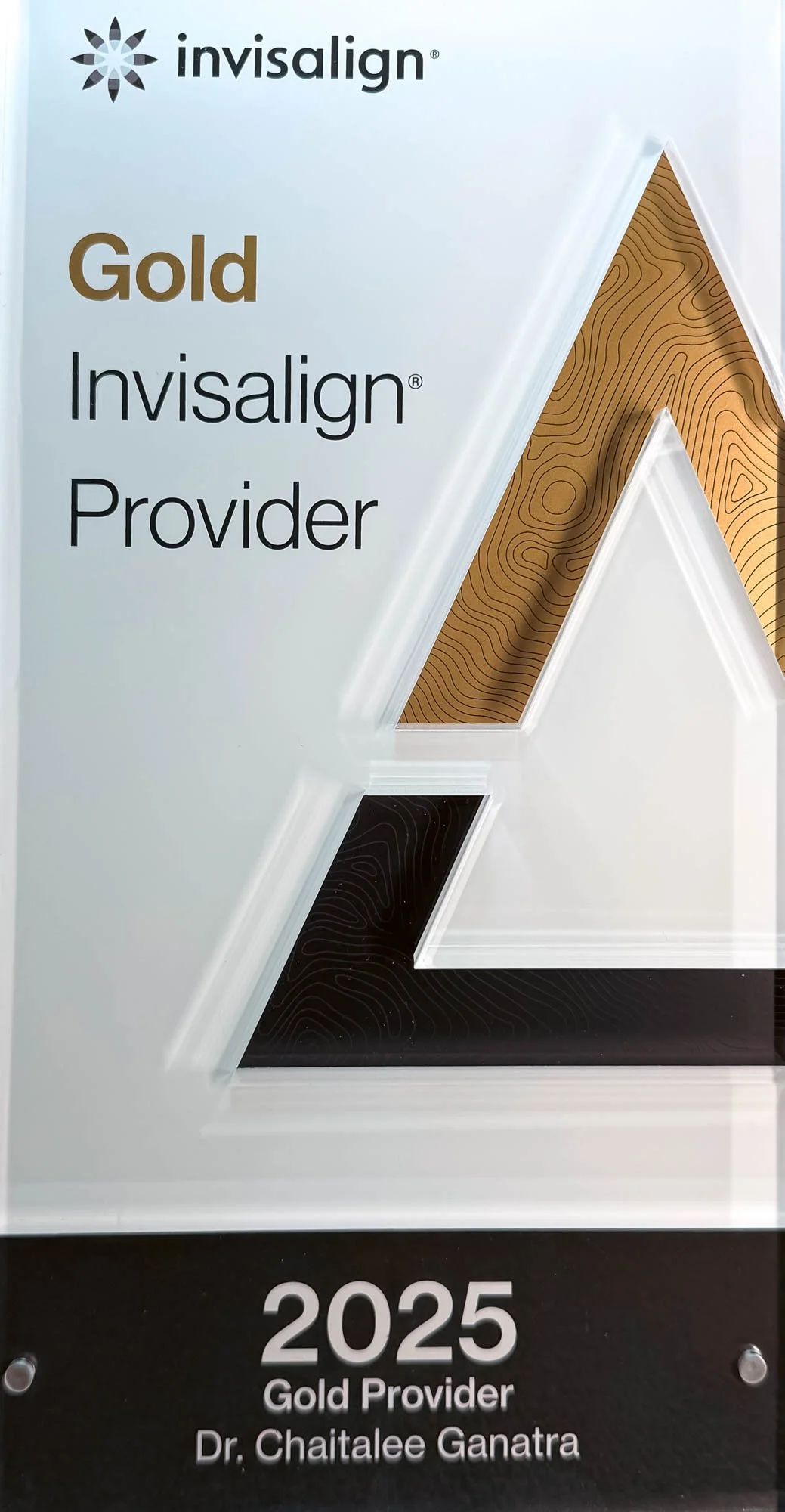 A plaque displaying Invisalign's 2025 Gold Provider award, awarded to Dr. Chaitalee Ganatra, with a large gold and black stylized 'A' logo design in the background.