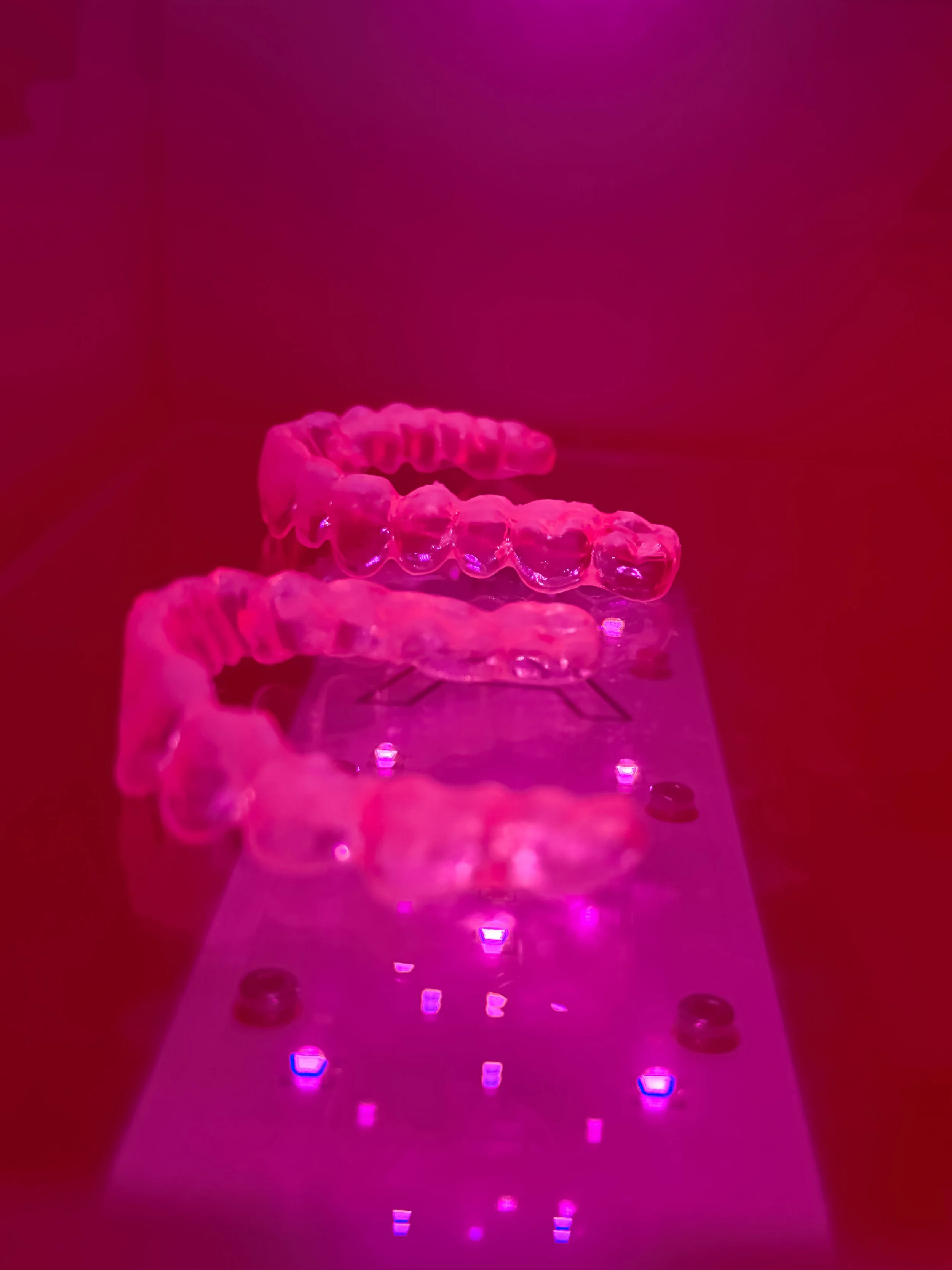 Clear dental aligners on a reflective pink surface with red lighting.