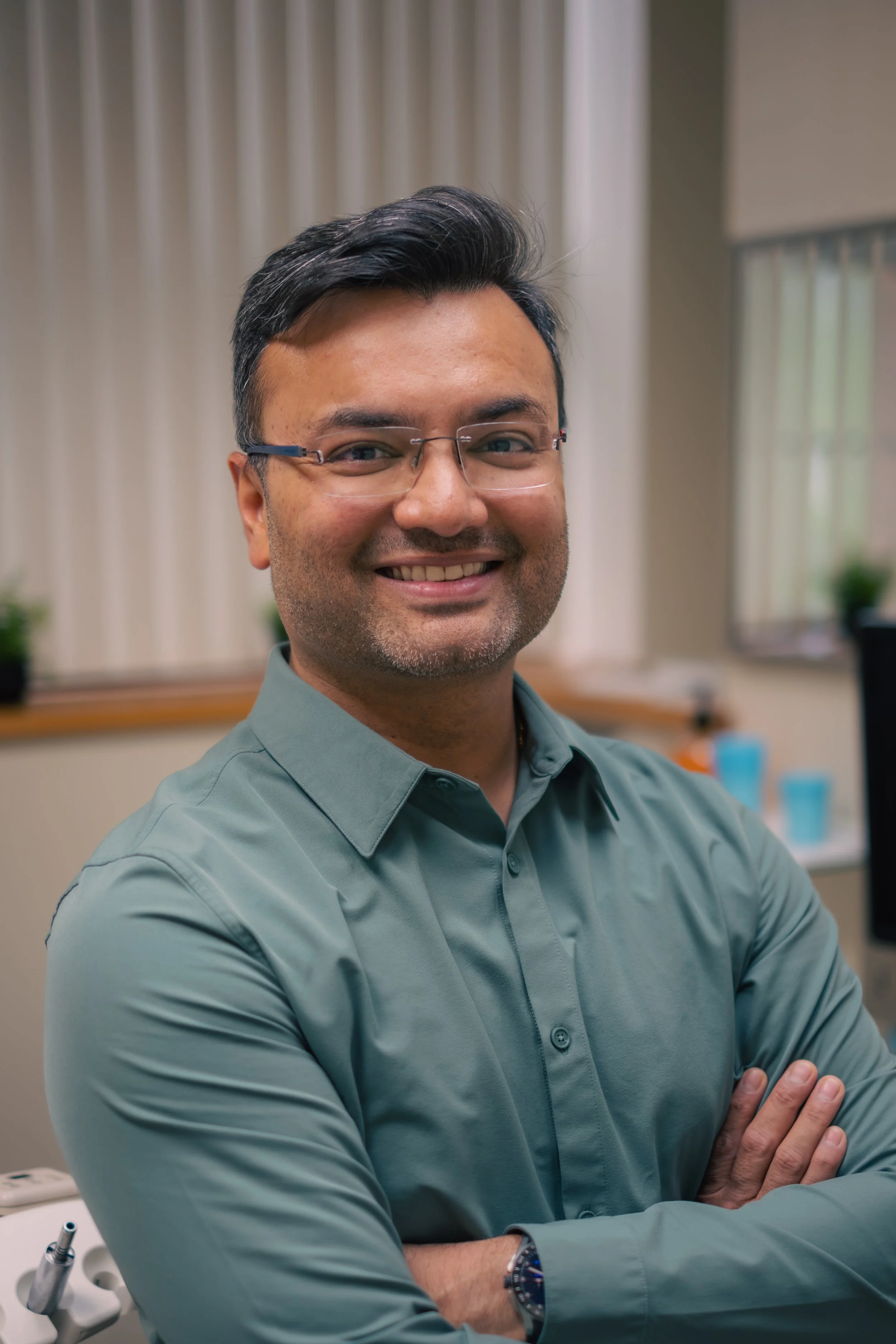 A man with glasses smiling, wearing a green shirt, with arms crossed, in a room with neutral curtains and some medical or office equipment in the background.