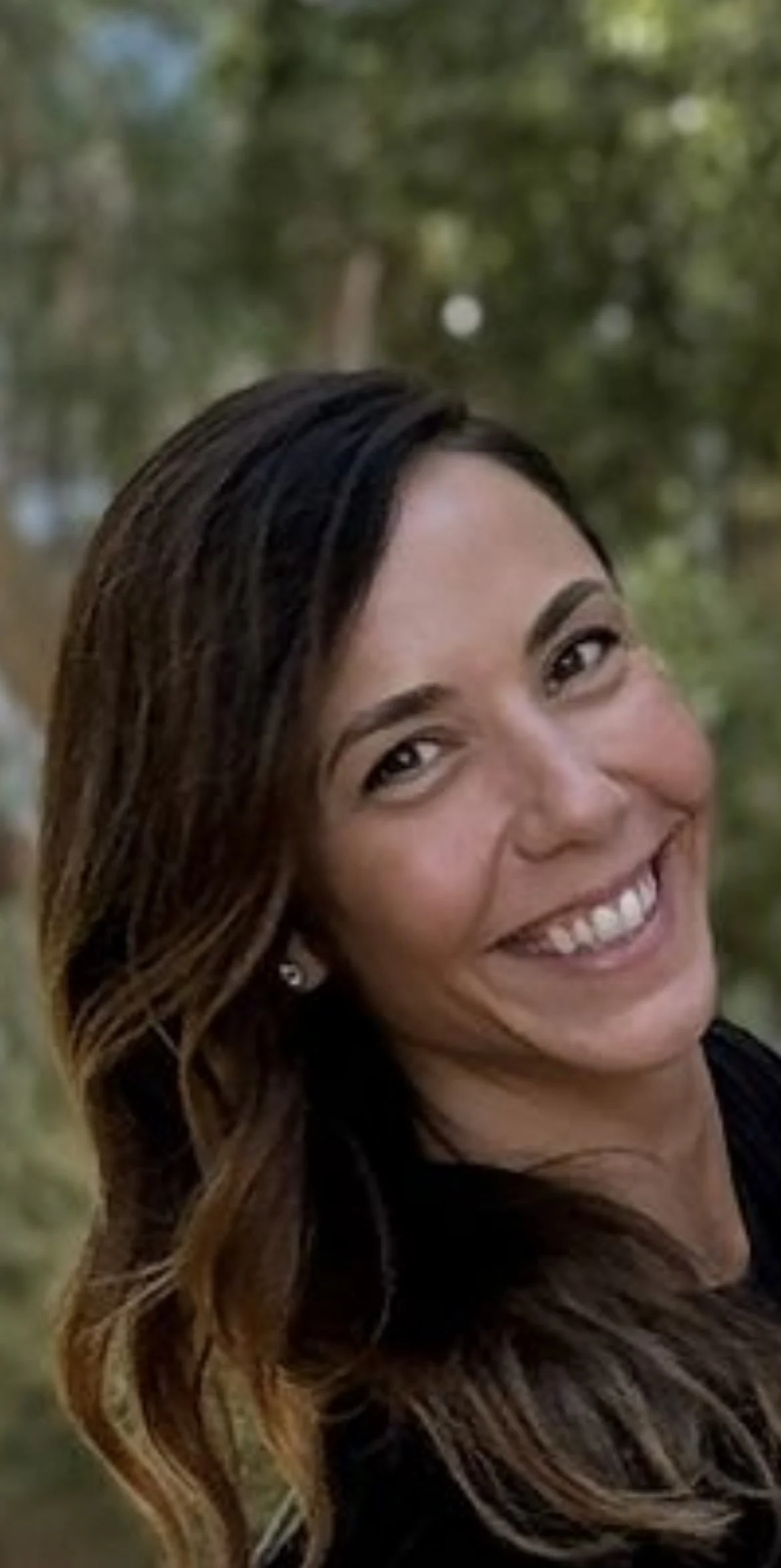 Close-up of a smiling woman with long, wavy brown hair, outdoors with blurred green trees in the background.