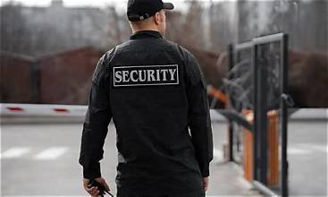 Security guard in black uniform with 'SECURITY' patch on back, walking away from camera near a gated area.