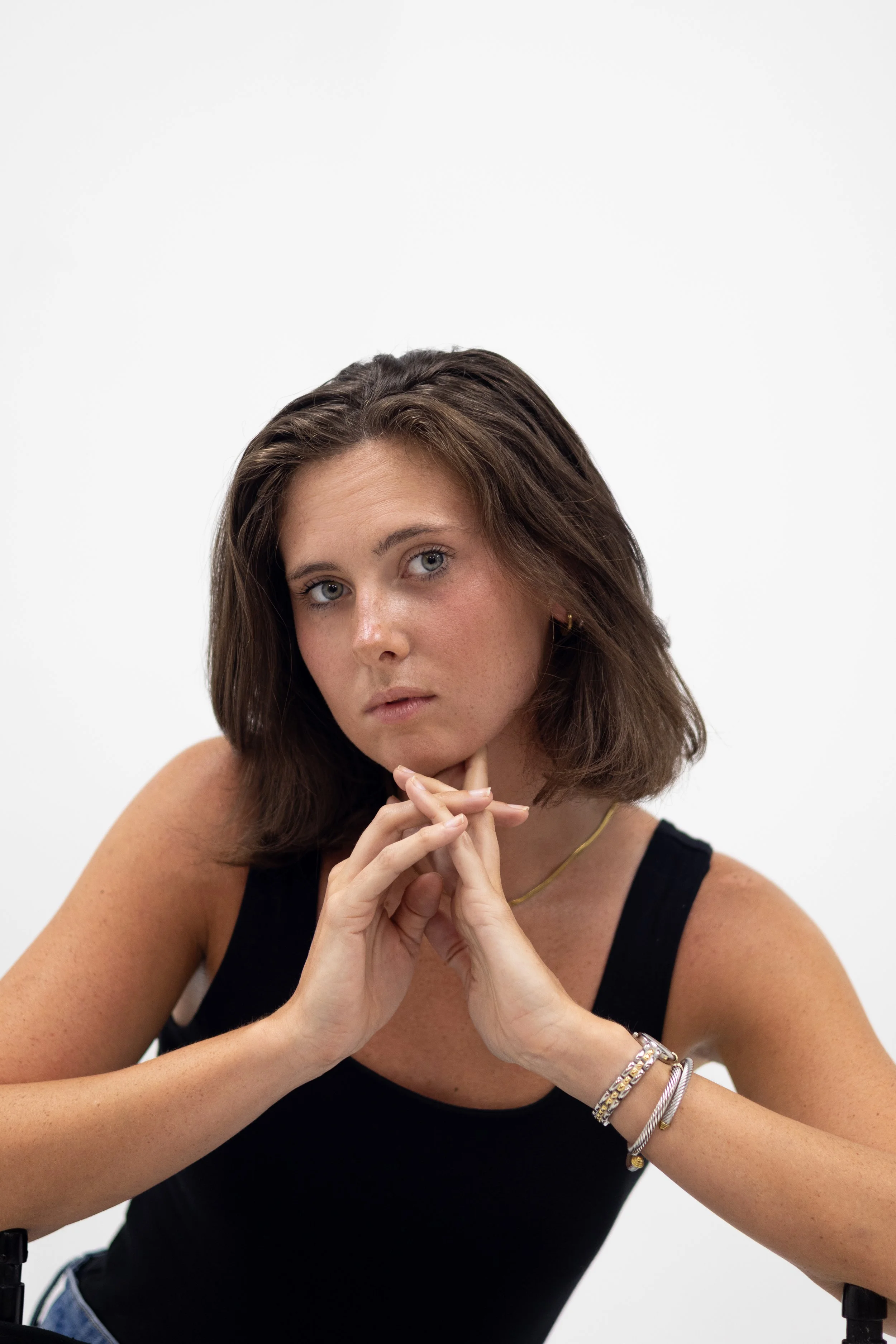 A young woman with shoulder-length brown hair, wearing a black sleeveless top, gold jewelry including a necklace and bracelets, is sitting against a plain white background. She has her hands clasped under her chin and is looking into the camera with 