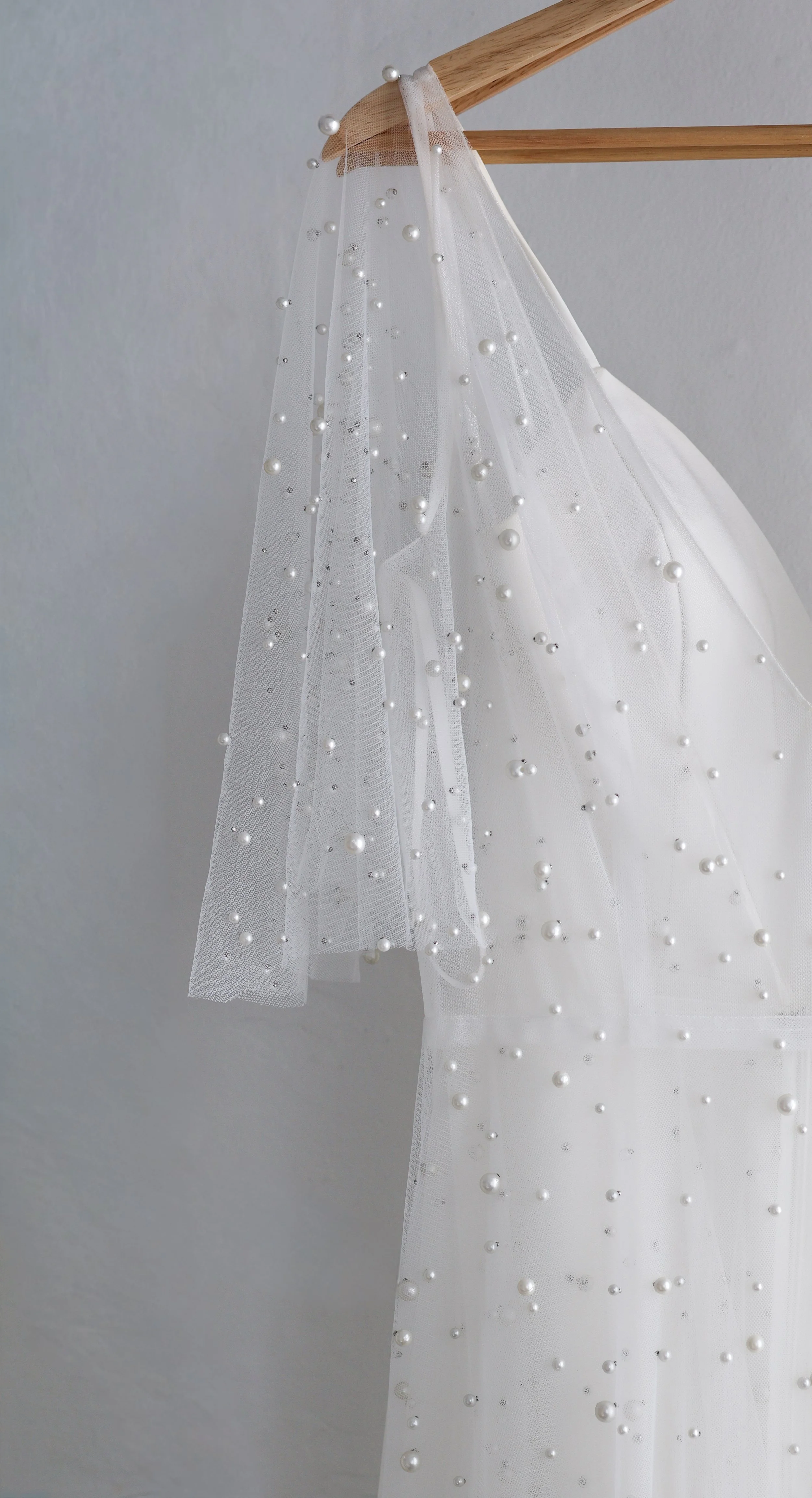 A white wedding veil with pearl embellishments hanging from a wooden hanger against a plain wall.