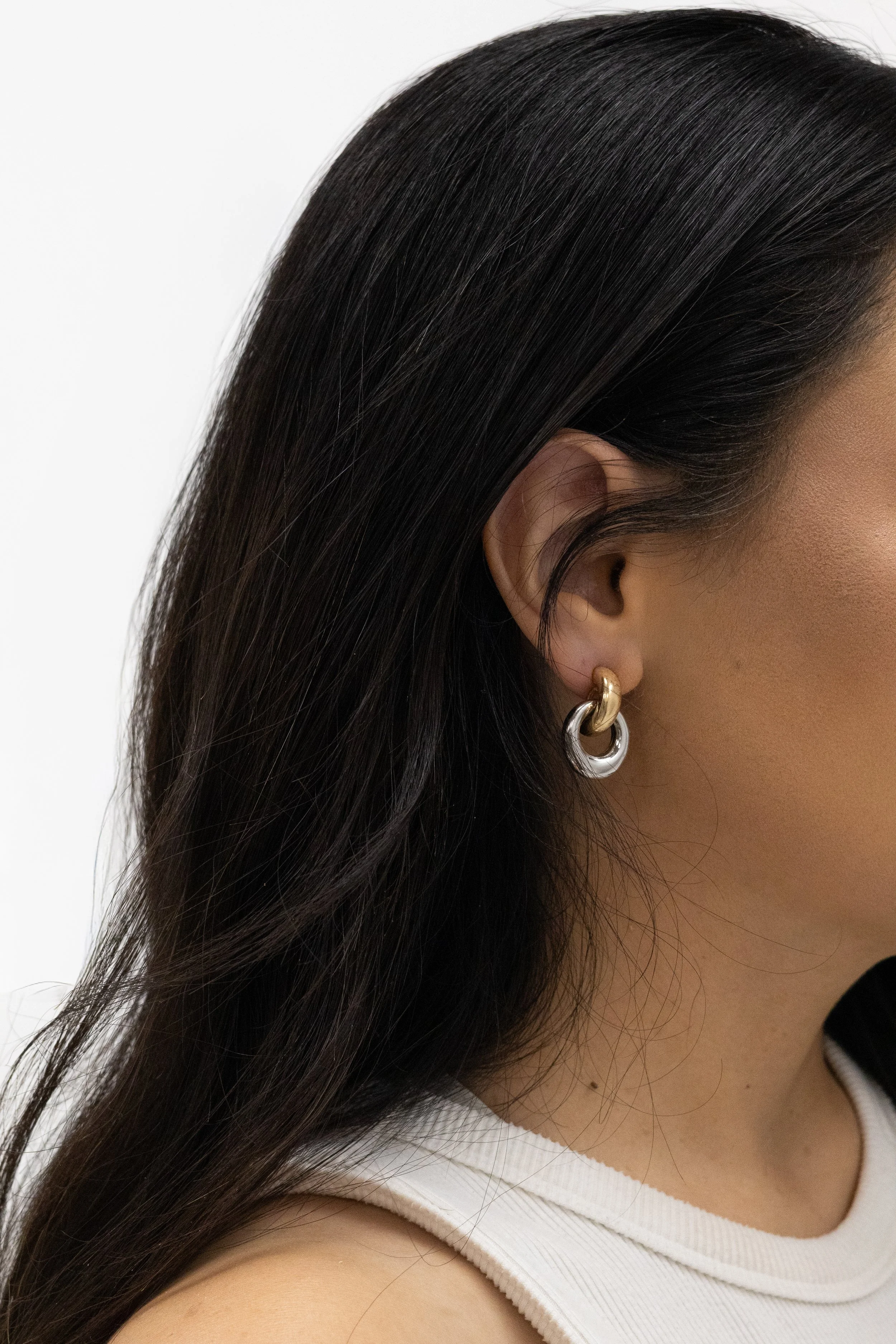 Close-up of a woman's face showing her dark hair, ear with gold and silver hoop earrings, and part of her shoulder in a white sleeveless top.