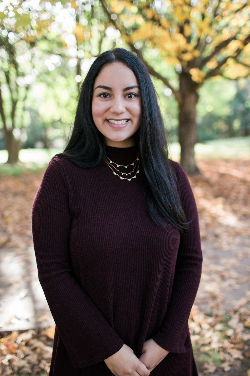 A young woman with long black hair, smiling, stands outdoors among trees with autumn-colored leaves, wearing a burgundy sweater and layered necklaces.