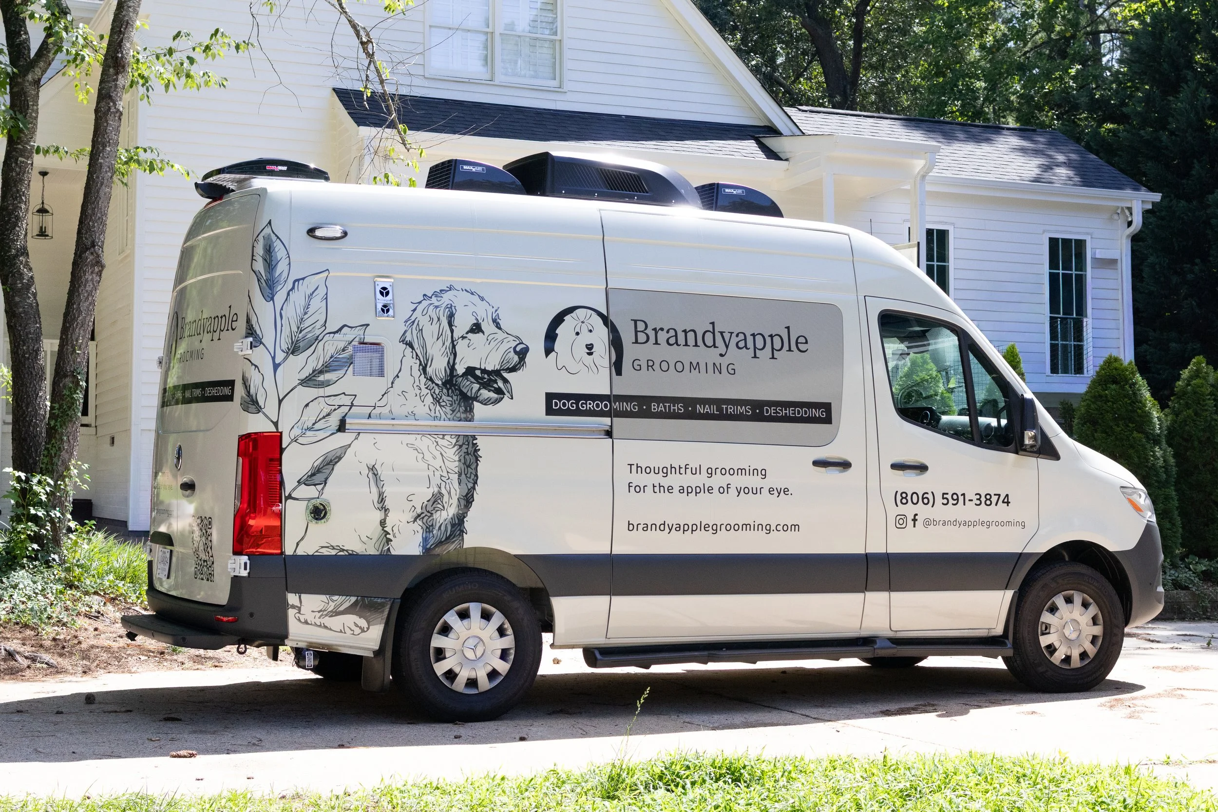 A white van with branding for Brandyapple Grooming, featuring dog illustrations and text about grooming services, parked in front of a white house with trees.