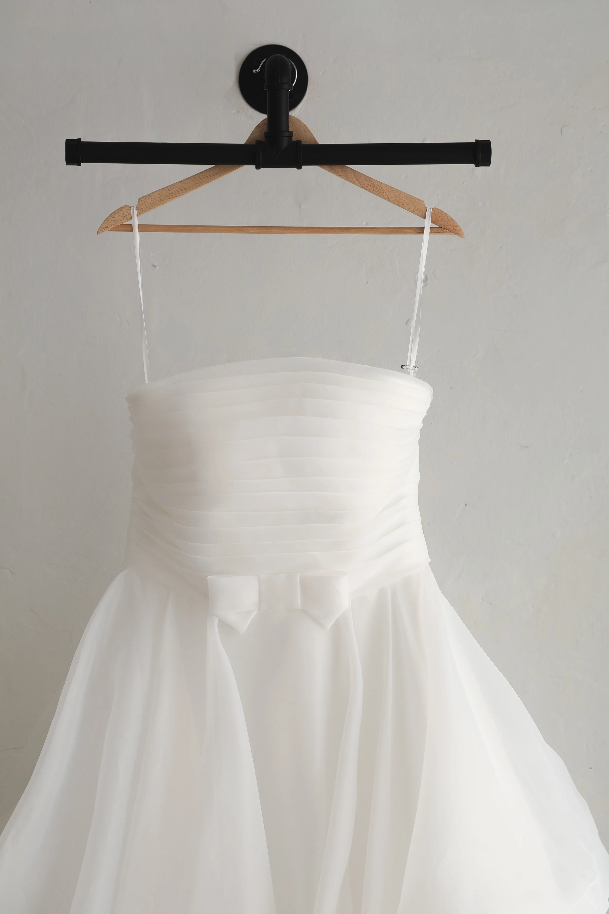 White wedding dress with pleated bodice and bow, hanging from a black pipe clothing rack attached to the ceiling.