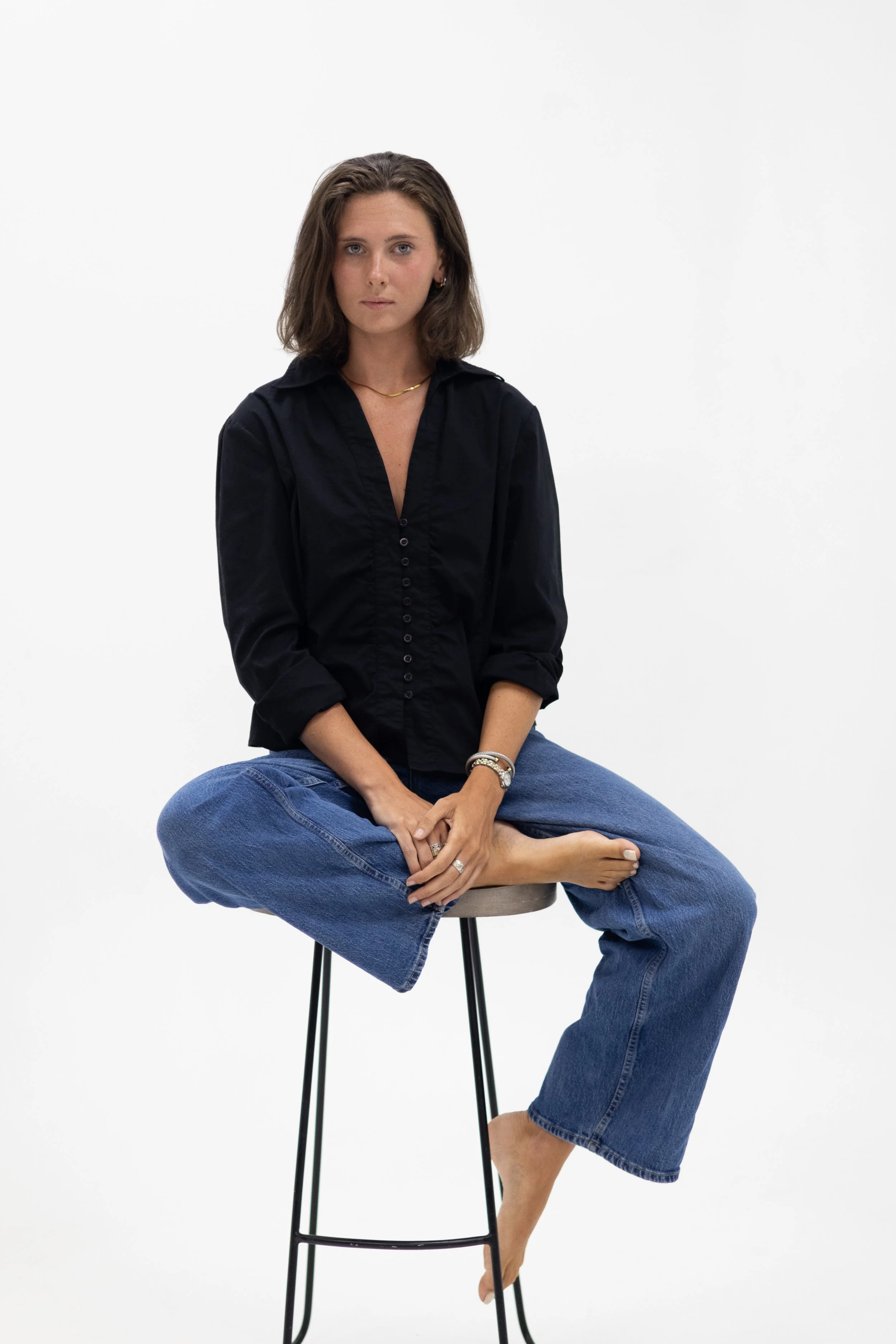 A woman with shoulder-length brown hair in a black shirt and blue jeans, sitting cross-legged on a stool against a plain white background.