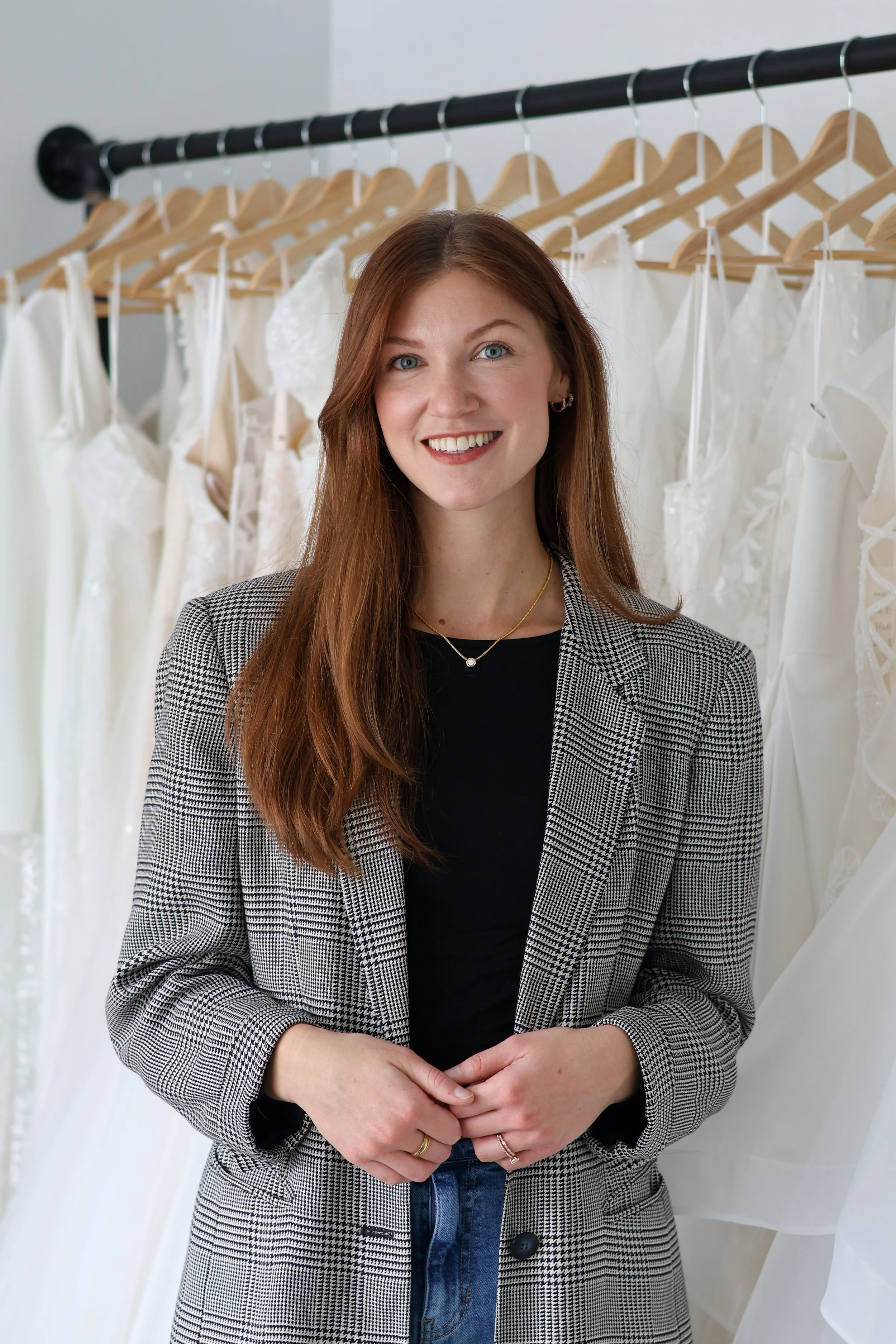 A smiling woman with long red hair wearing a black top and a checkered blazer, standing in front of a clothing rack with white dresses on wooden hangers.