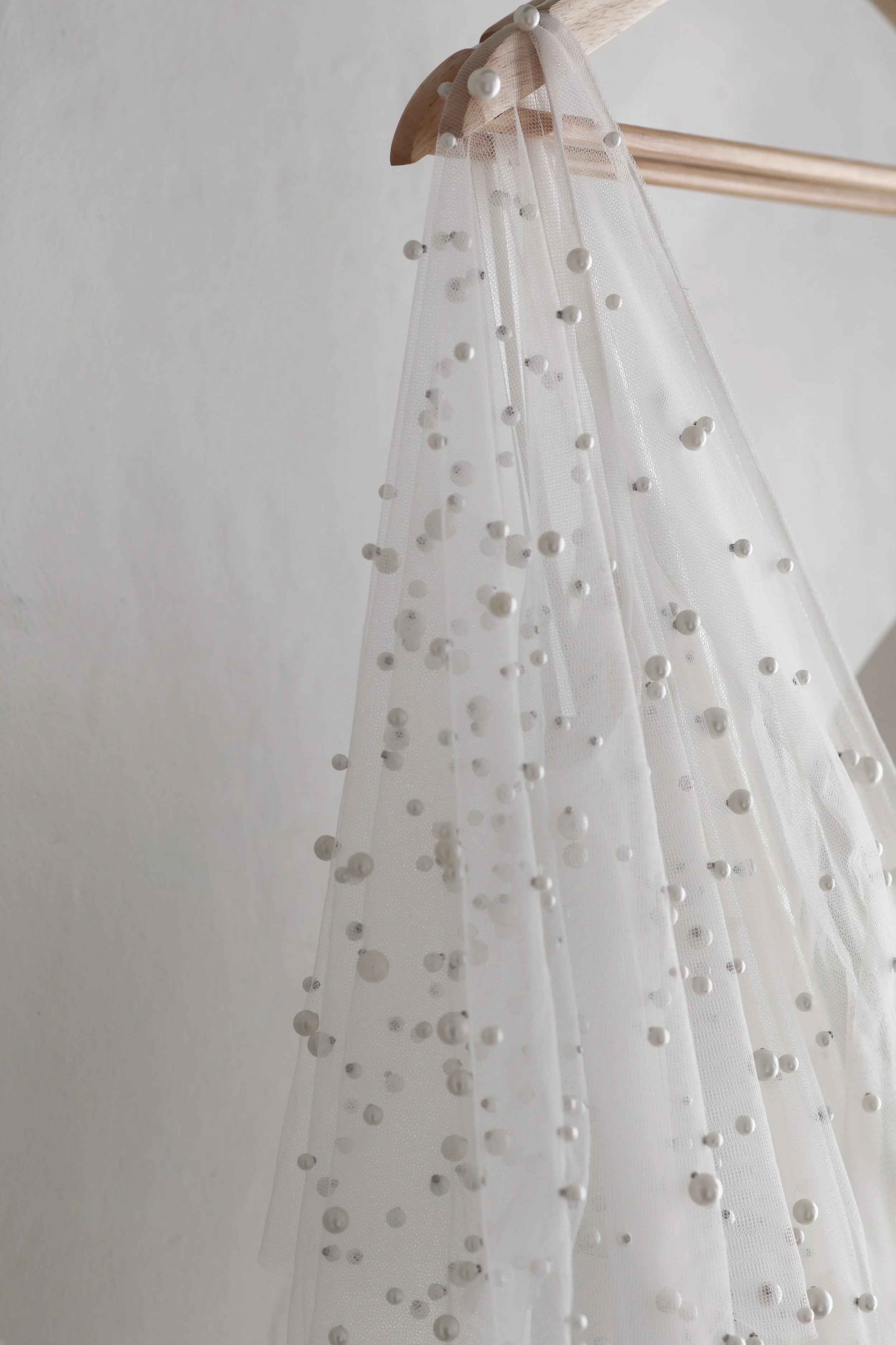 A white, transparent fabric with pearl-like beads hanging from a wooden hanger against a white wall.