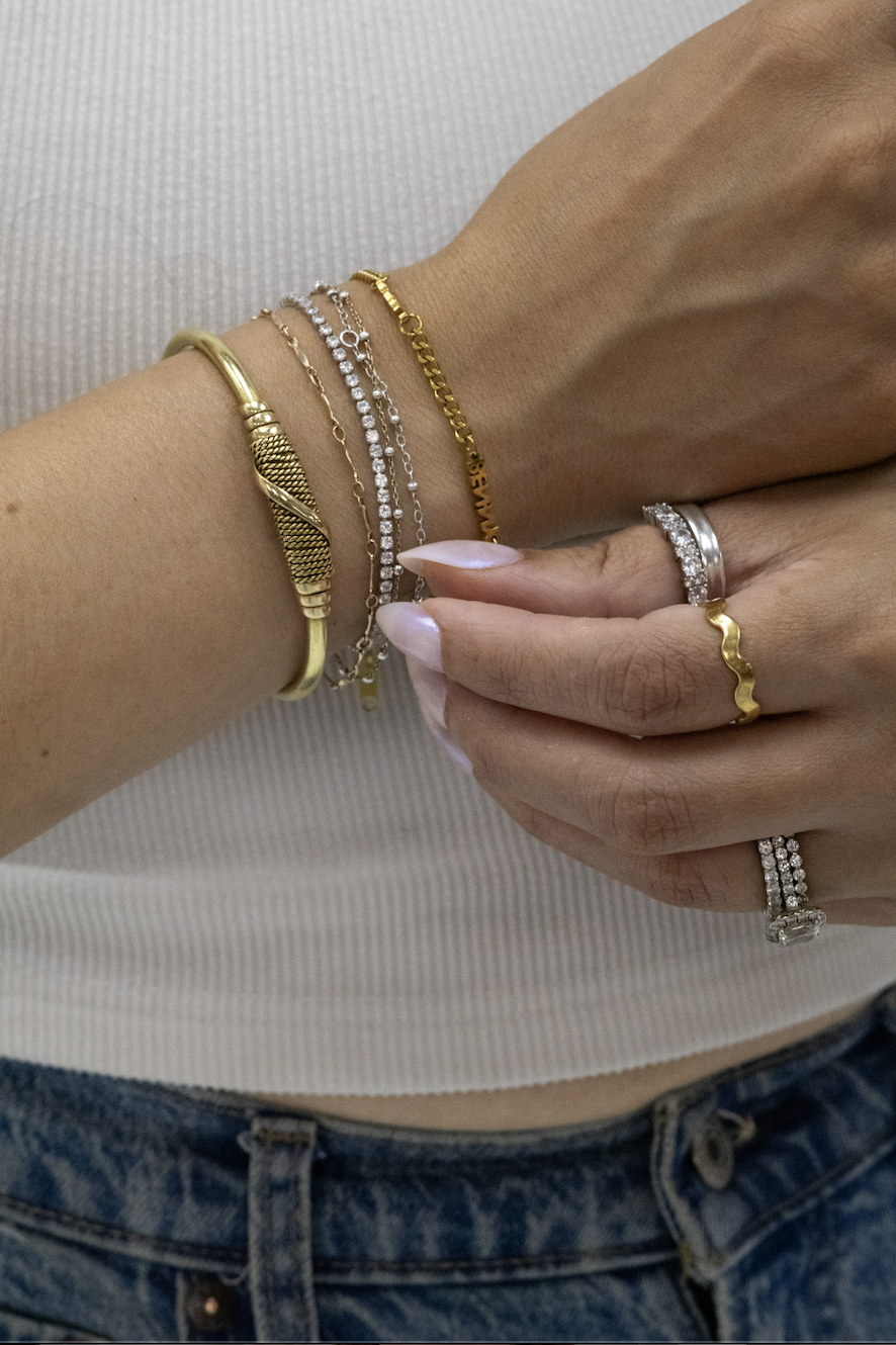A person's hand and wrist showing multiple gold and silver rings and bracelets with a white top and blue jeans.