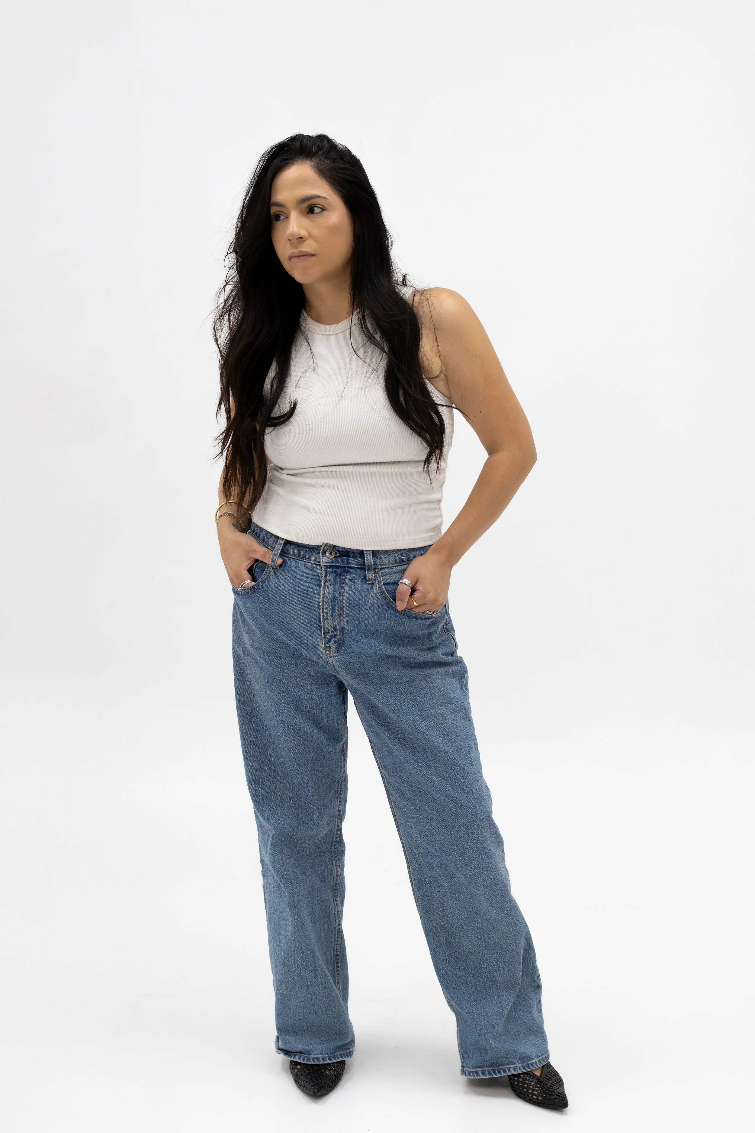 Woman standing with hands in pockets, wearing a white sleeveless top, high-waisted jeans, and black shoes, against a plain white background.