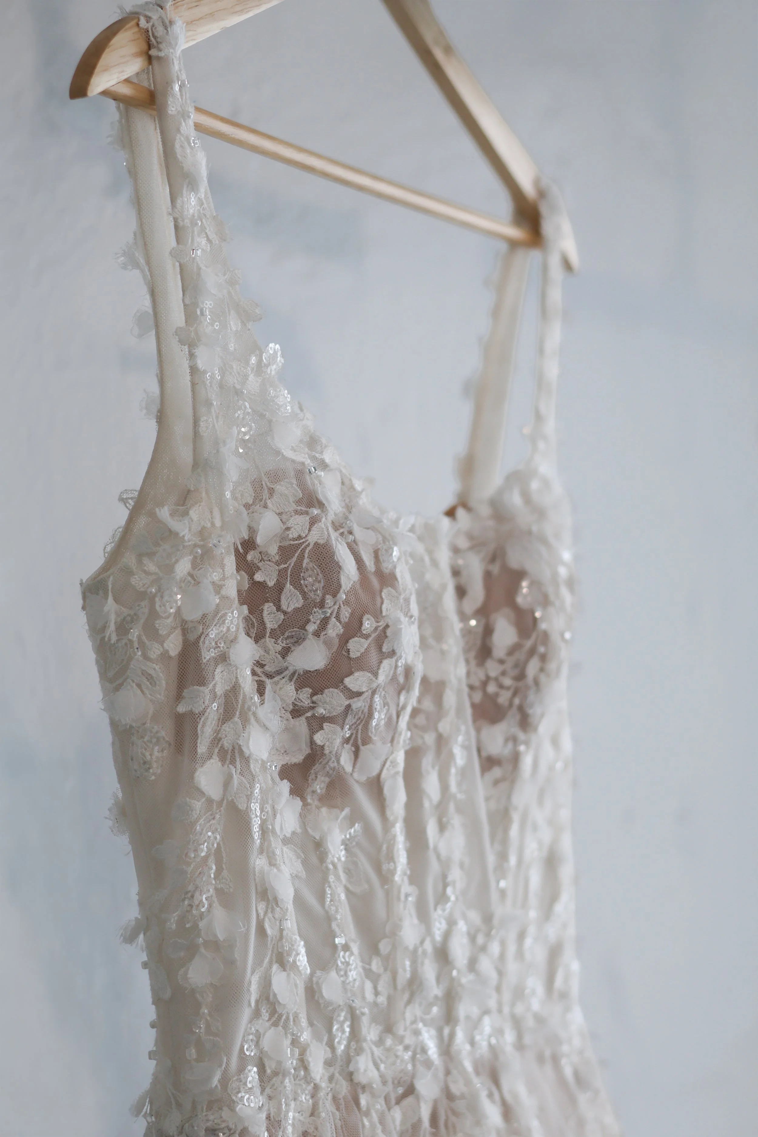 A delicate, cream-colored lace dress with floral embroidery and sequins hanging on a wooden hanger against a plain wall.