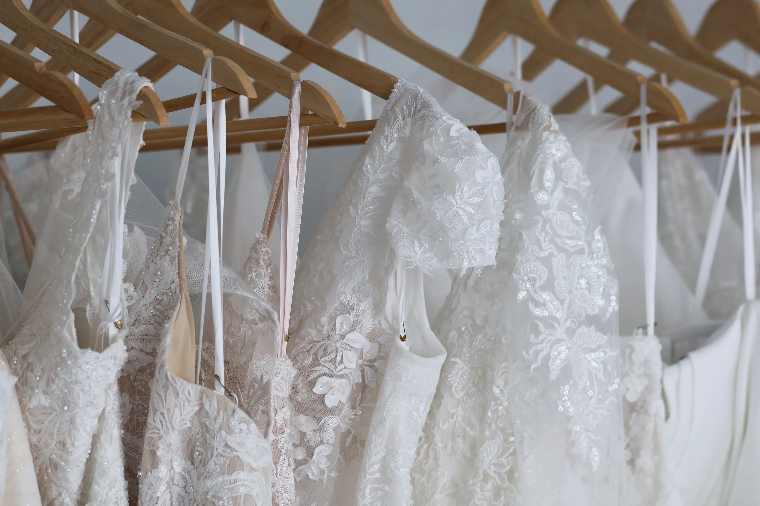A row of white lace wedding dresses hanging on wooden hangers on a wooden clothing rack.
