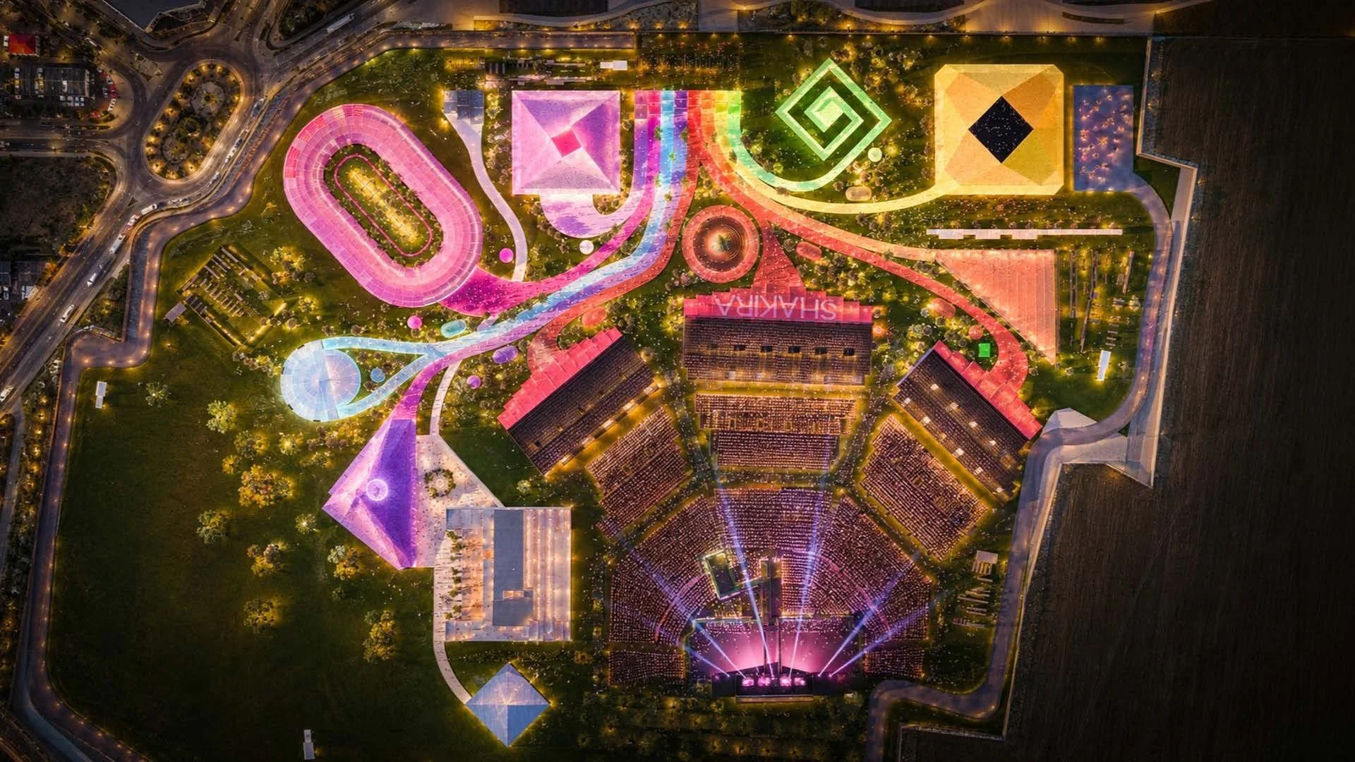 Vista aérea nocturna de Estadio Shakira con luces de diferentes colores, incluyendo una pista de carreras, un escenario con espectáculo y varios edificios y zonas de juego iluminadas.