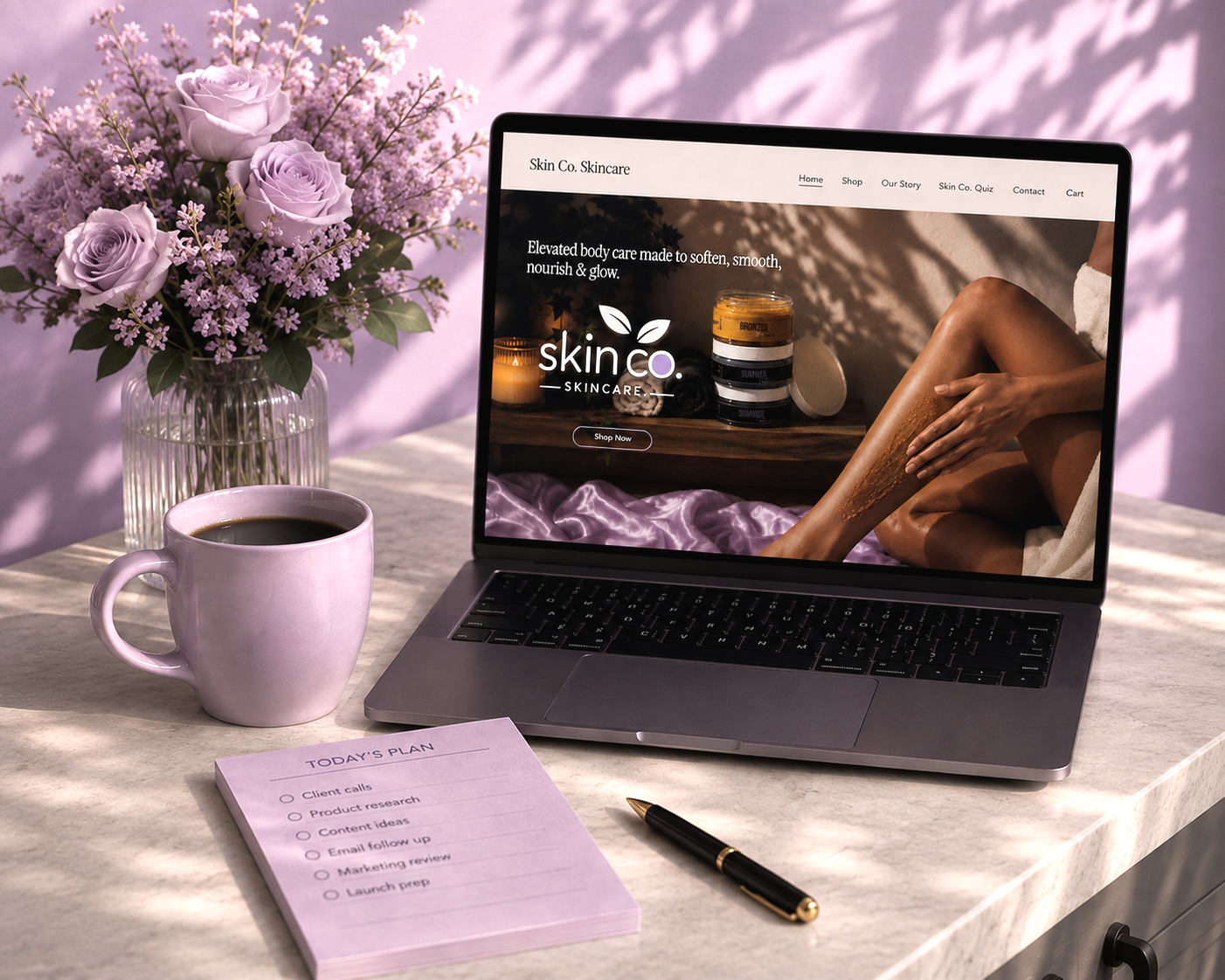 Workspace with a laptop displaying the Skin Co. website, flowers, a coffee mug, and a notepad.