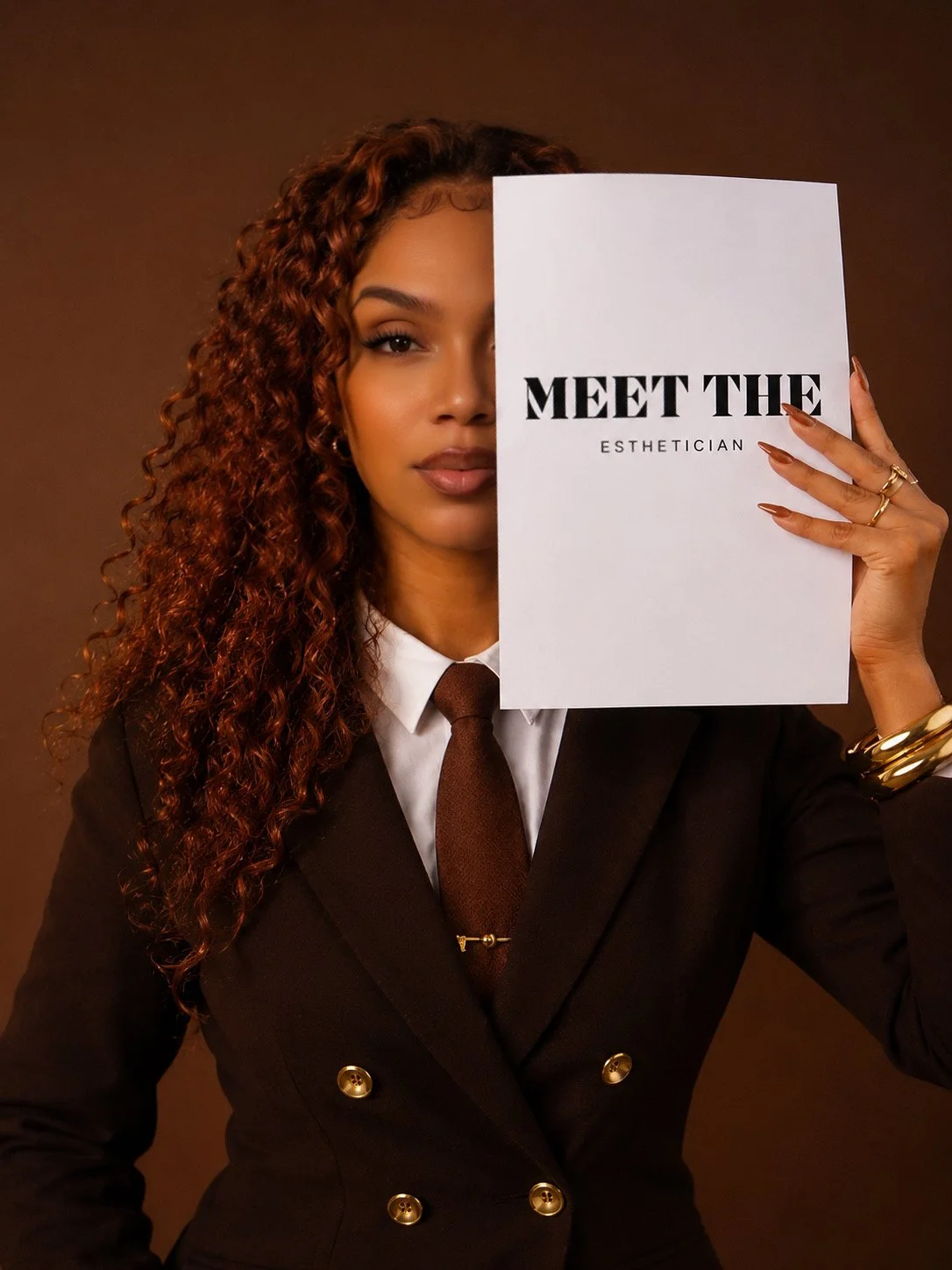 A woman with curly hair dressed in a black suit with gold buttons, holding a white sign that says 'Meet the Esthetician' in front of her face.