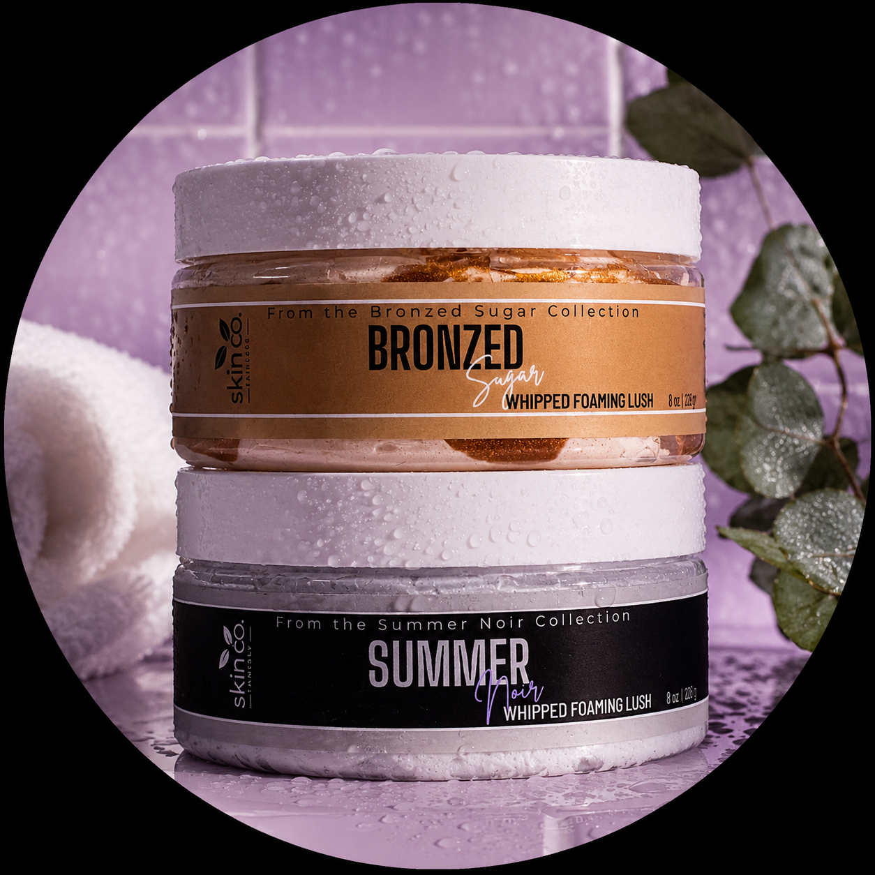 Stacked jars of skincare products with water droplets on them, labeled 'Bronzed Sugar Whipped Foaming Lush' and 'Summer Noir Whipped Foaming Lush', against a purple tiled background with green leaves on the side.
