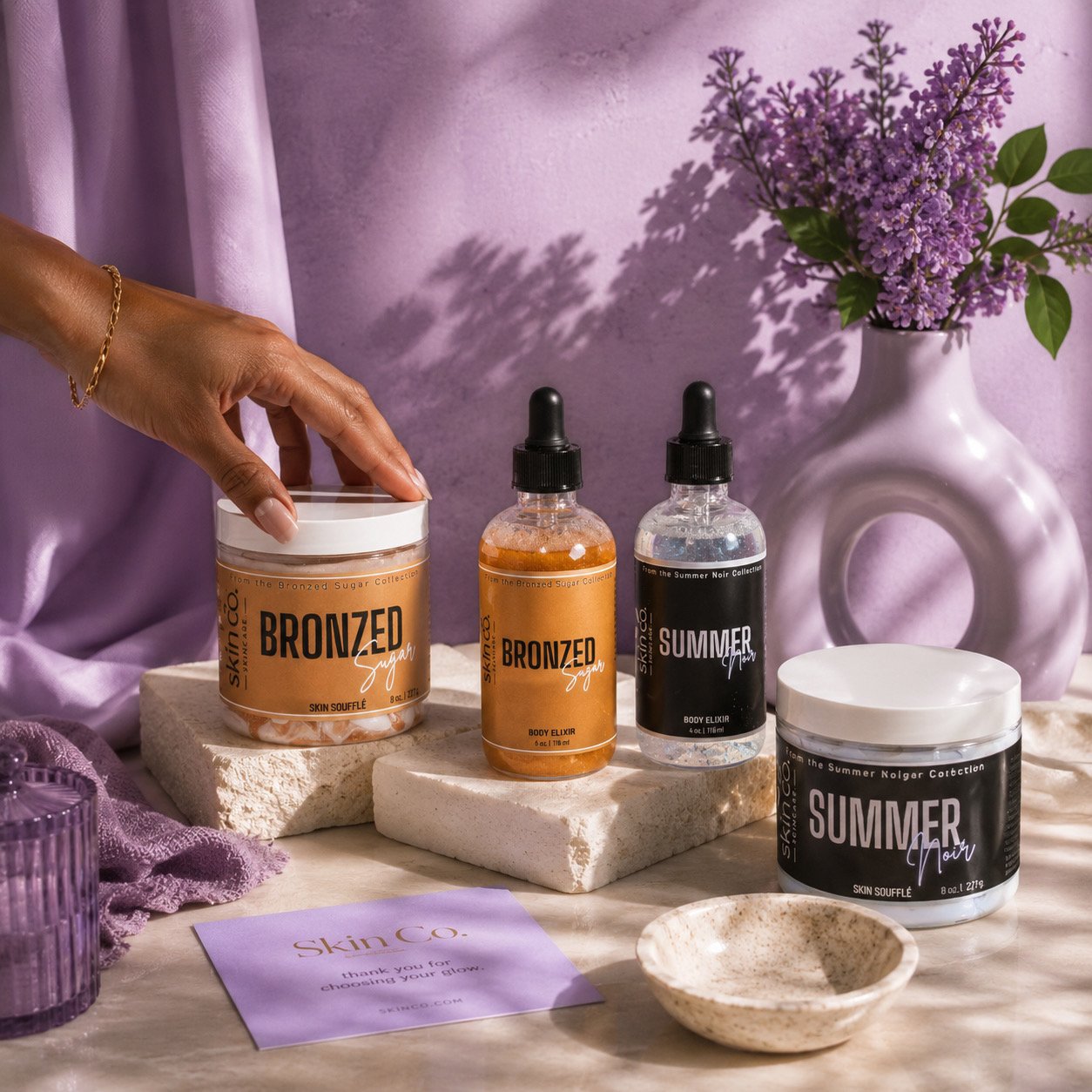 Bronzed Sugar and Summer Noir body care products arranged on marble counter with purple flowers.