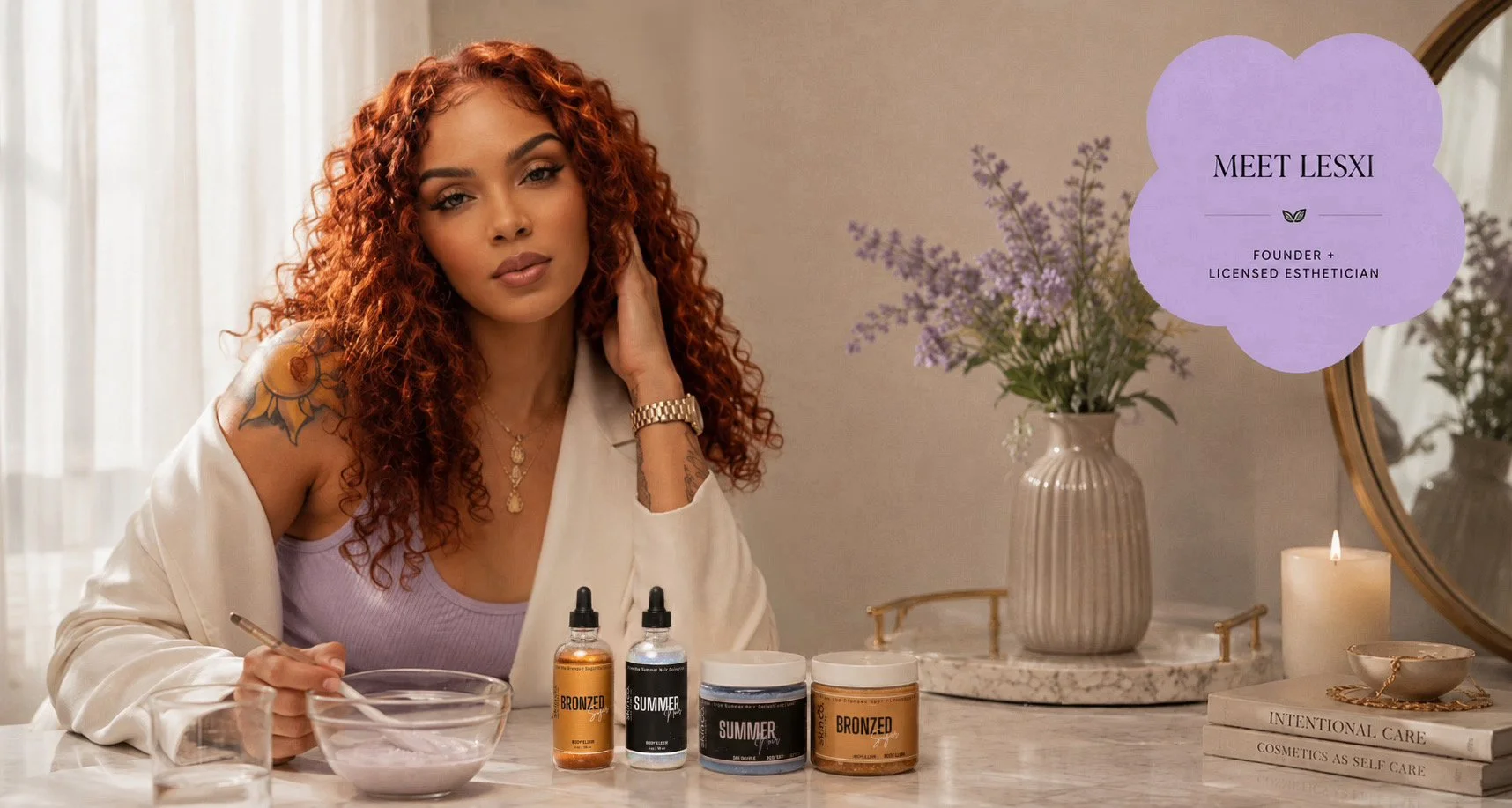 A woman with curly red hair and tattoos on her shoulder and arm sits at a marble vanity with skincare products, a glass bowl, and glasses of water. She is wearing a white blazer and purple top. Behind her, there is a vase with purple flowers and a lit candle, along with a mirror and two books, one titled 'Intentional' and the other 'Cosmetics as Self Care.' A purple speech bubble reads 'Meet Lexi, founder and licensed esthetician.'